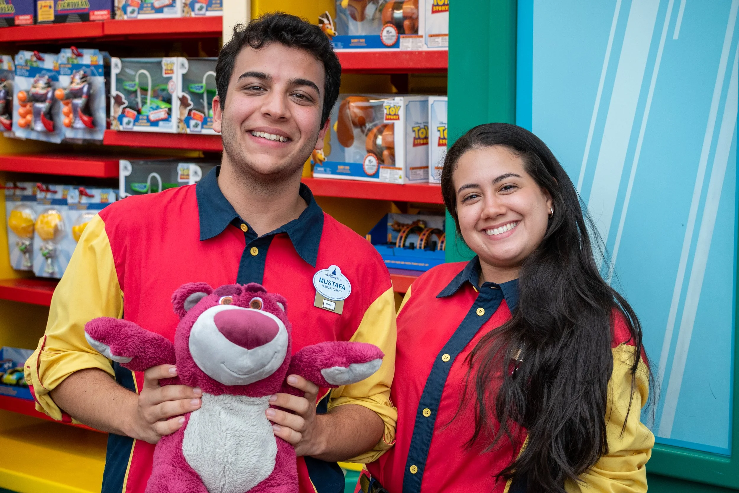 Two employees in red and yellow uniforms smiling while holding a pink and white plush toy lion inside a toy store.