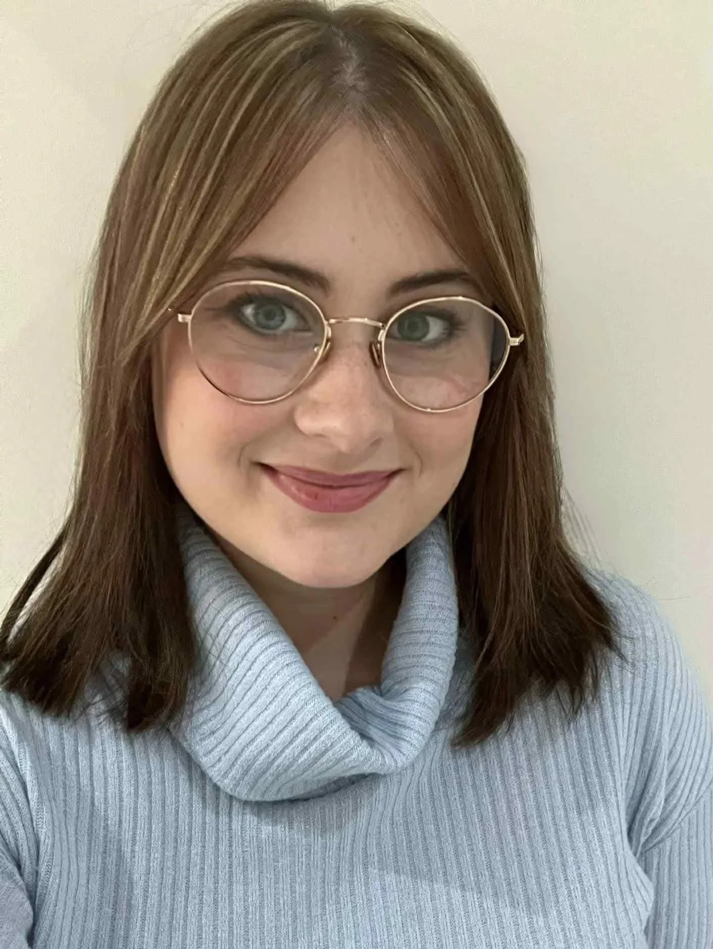 A woman with shoulder-length reddish-brown hair, wearing round glasses with a gold frame, a light gray turtleneck sweater, and smiling at the camera against a plain light-colored background.