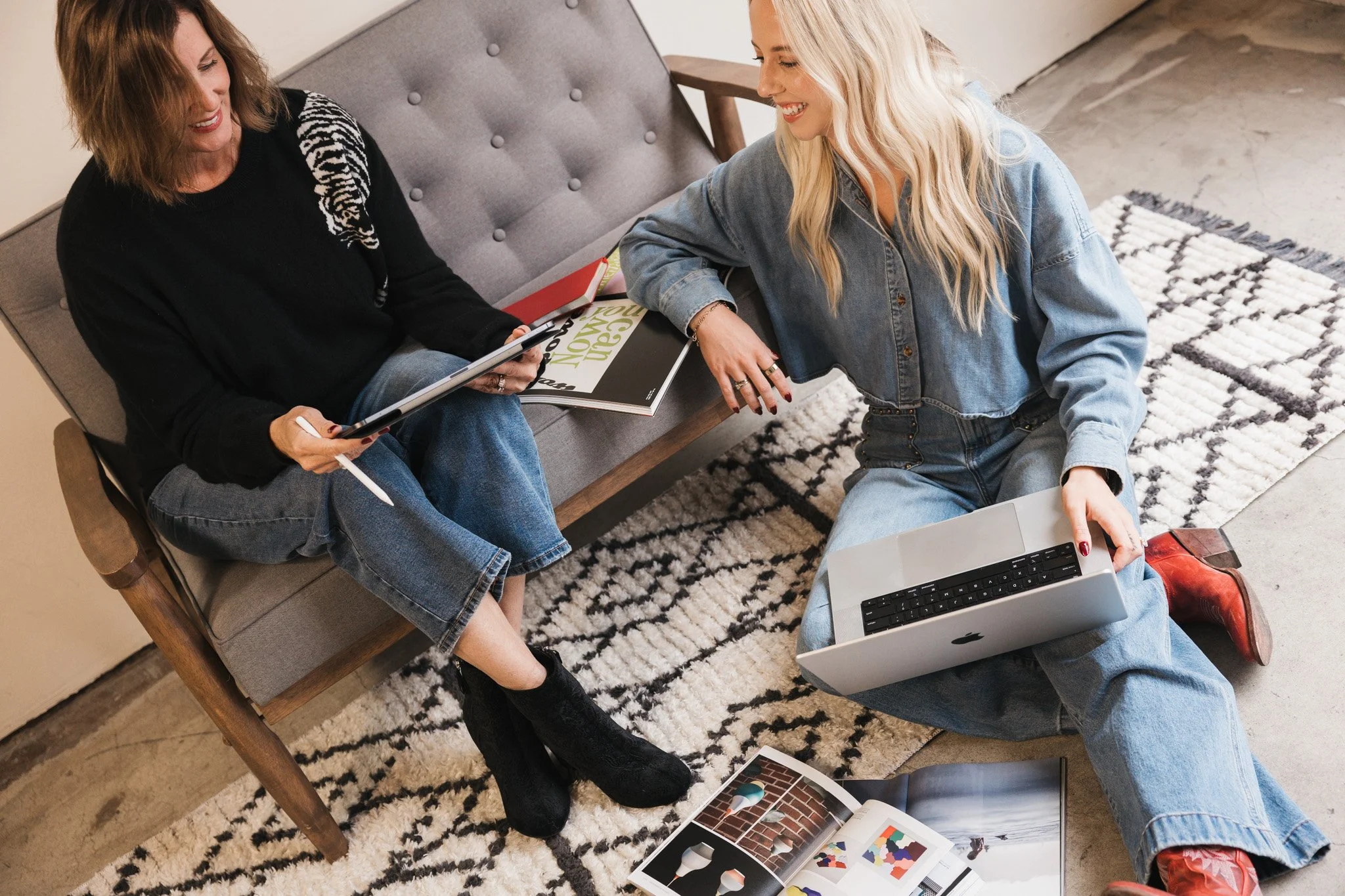 Two women sitting on a beige couch and on the floor, smiling and talking. One woman is holding a tablet, and the other is using a laptop. There are books, magazines, and photographs on the floor and on the couch.