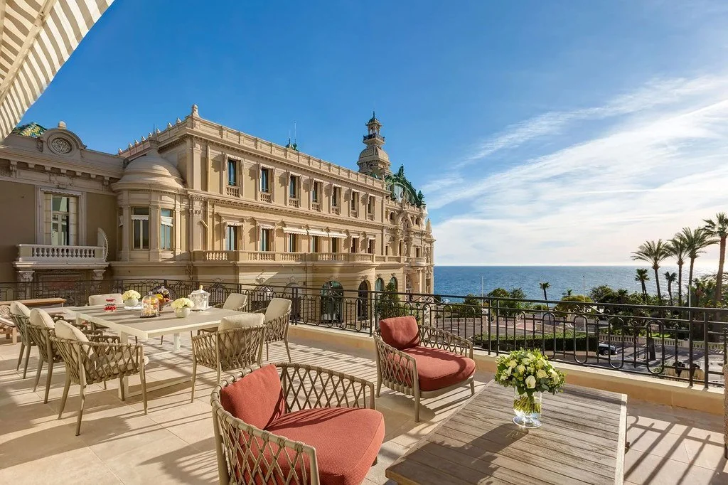 Luxury balcony with dining table, seating, and flowering centerpiece, overlooking the ocean and historic hotel building with palm trees in the background.