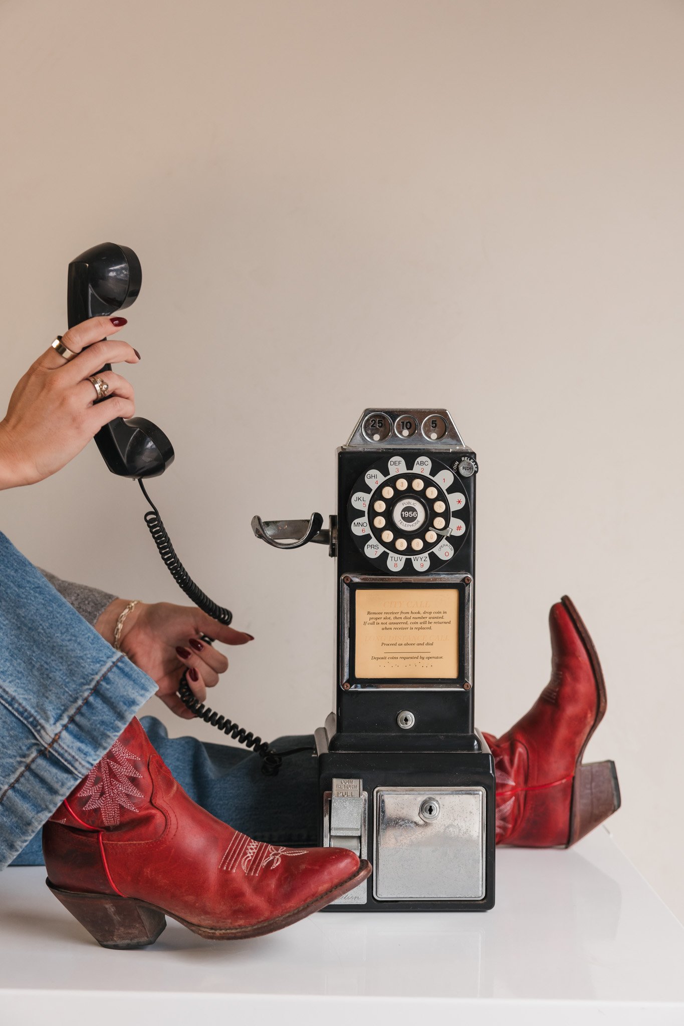 Person wearing red cowboy boots, sitting at a white table and holding an old-fashioned black rotary dial telephone, with a vintage black payphone in front of them.