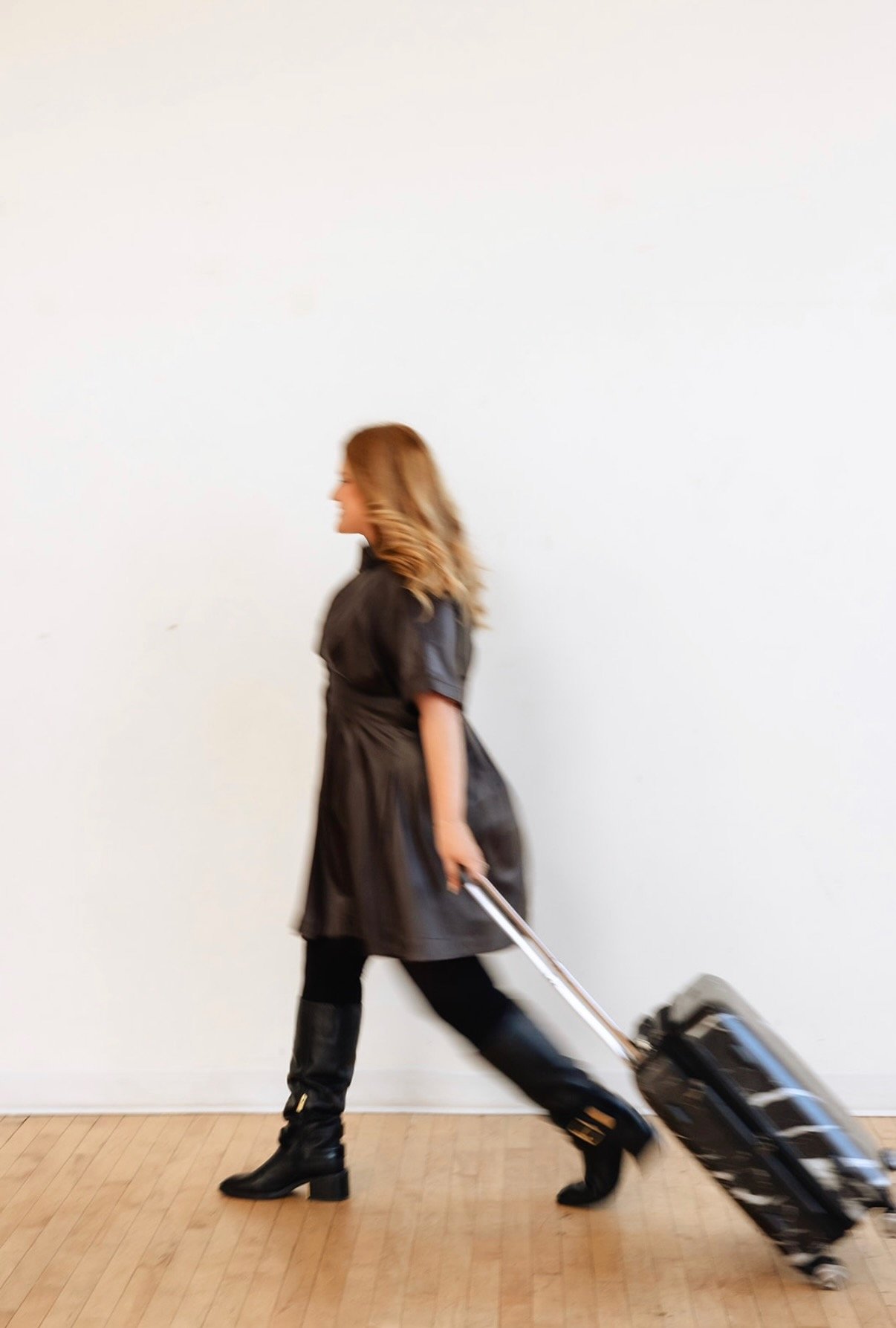 Kelli, from Let Your Mind Unwind, in black boots and a black dress walks while pulling a small rolling suitcase with a black and white pattern. Luxury travel advisor based in San Diego.