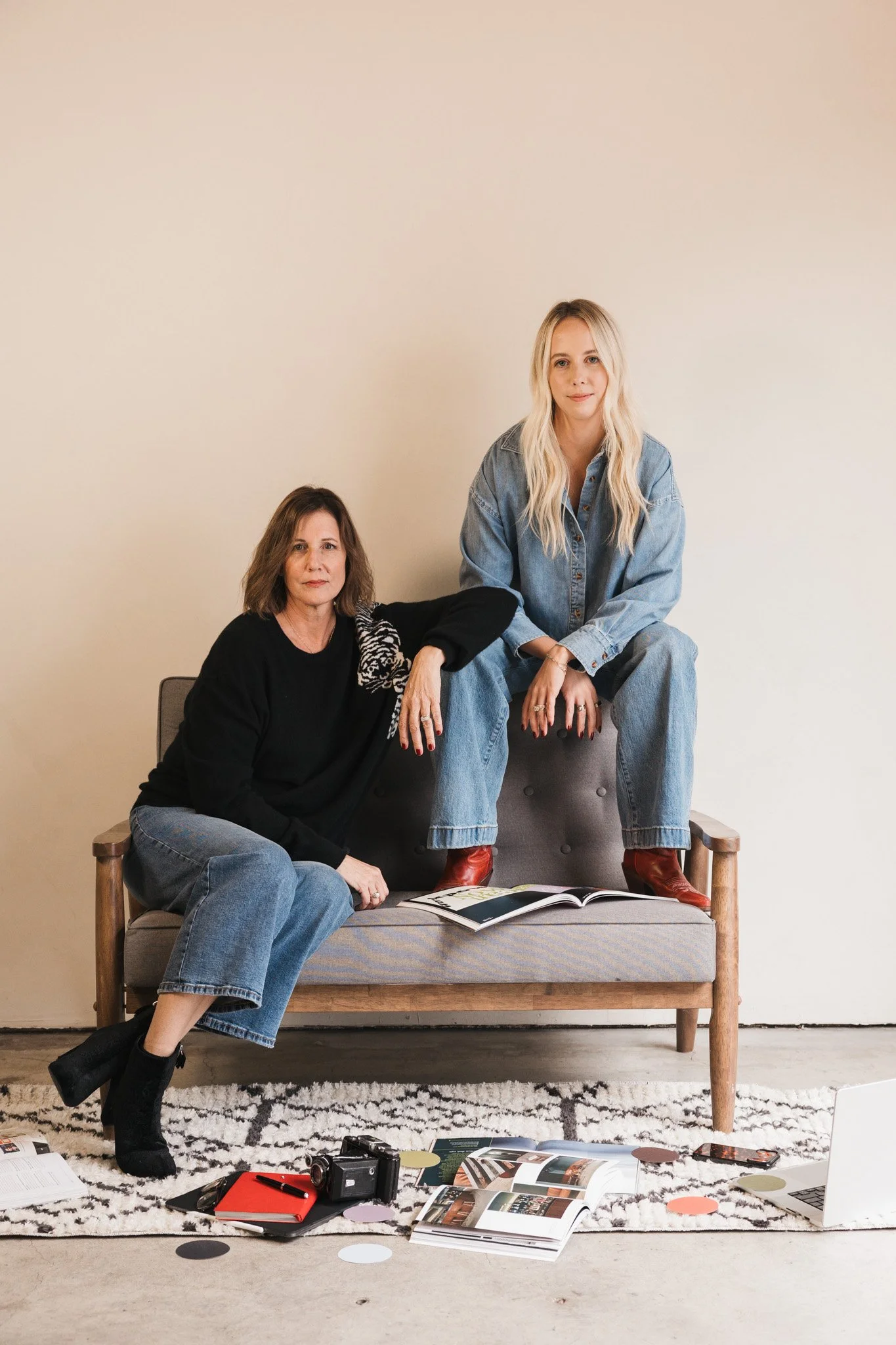 Two women sit on and around a sofa in a room with a light-colored wall; one is sitting on the sofa with her legs crossed, and the other is sitting on the armrest of the sofa, with an open magazine on her lap. Carson and April from Adverb Collective.
