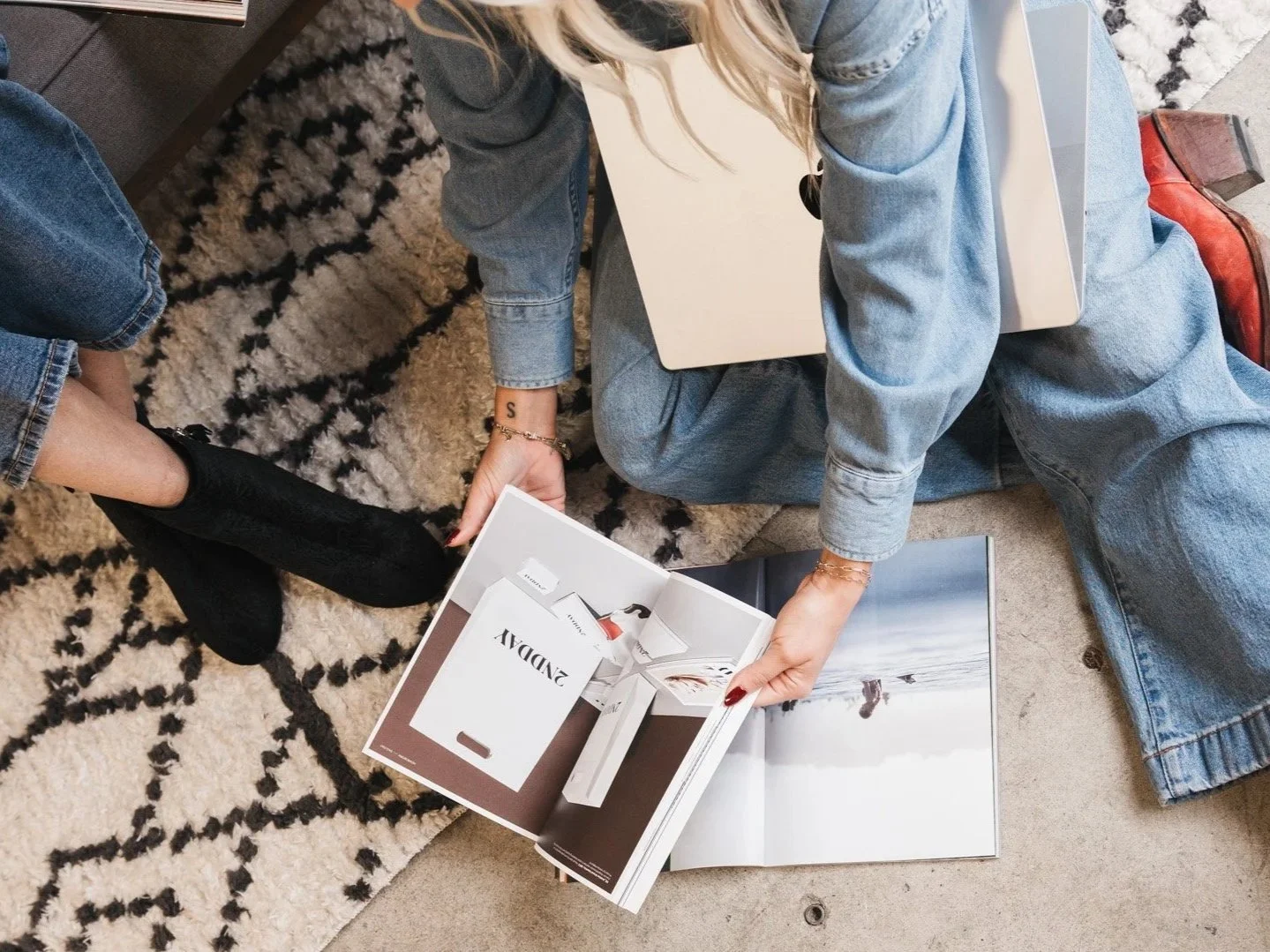 Two women sitting on the floor, looking at a magazine, with one woman holding it open to show a product advertisement. One woman is wearing a denim jacket, ripped jeans, and black boots, while the other is wearing a light denim outfit and red boots, with a laptop on her lap.