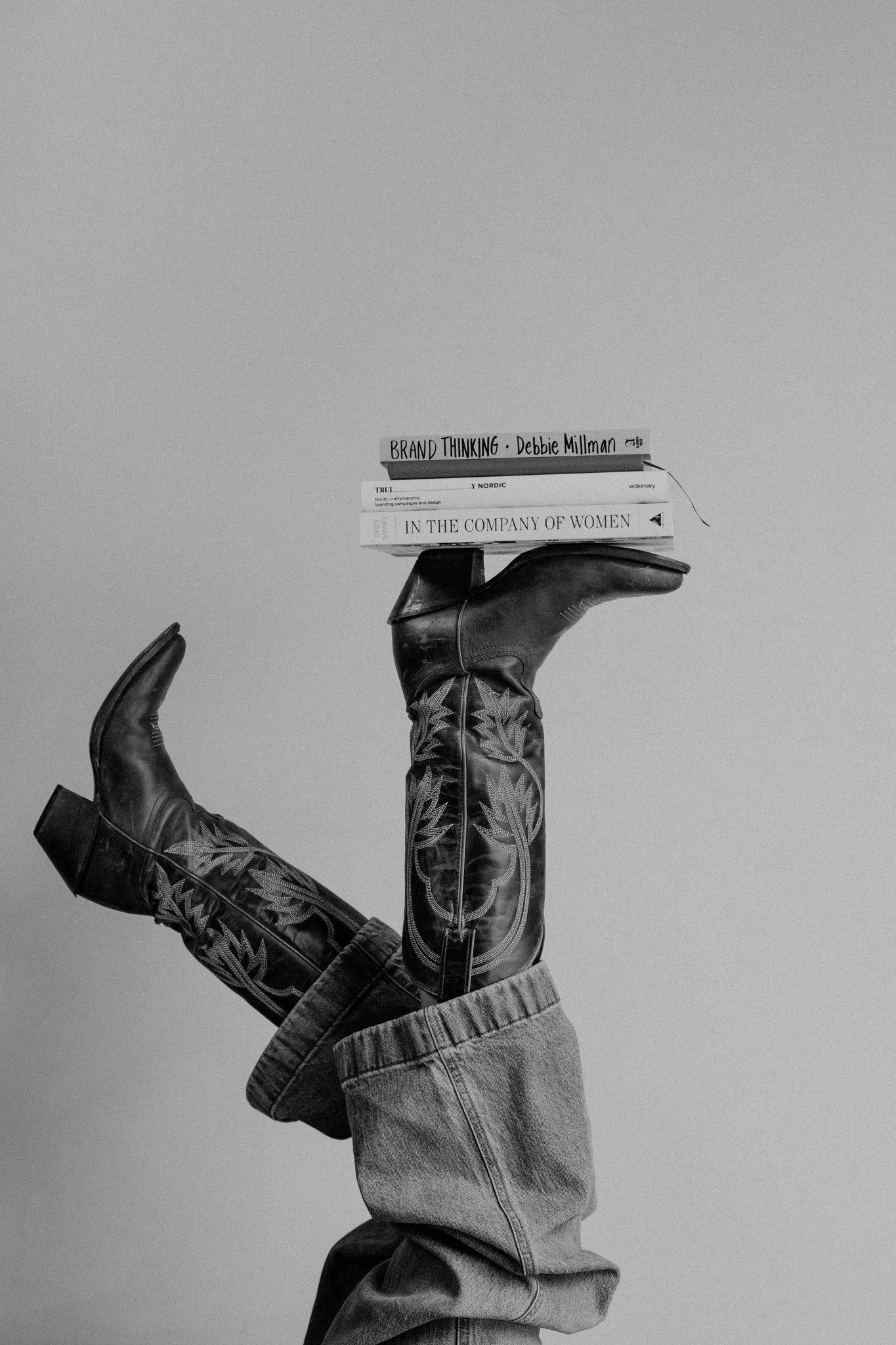 Person wearing jeans and cowboy boots, holding several books on their foot against plain background.