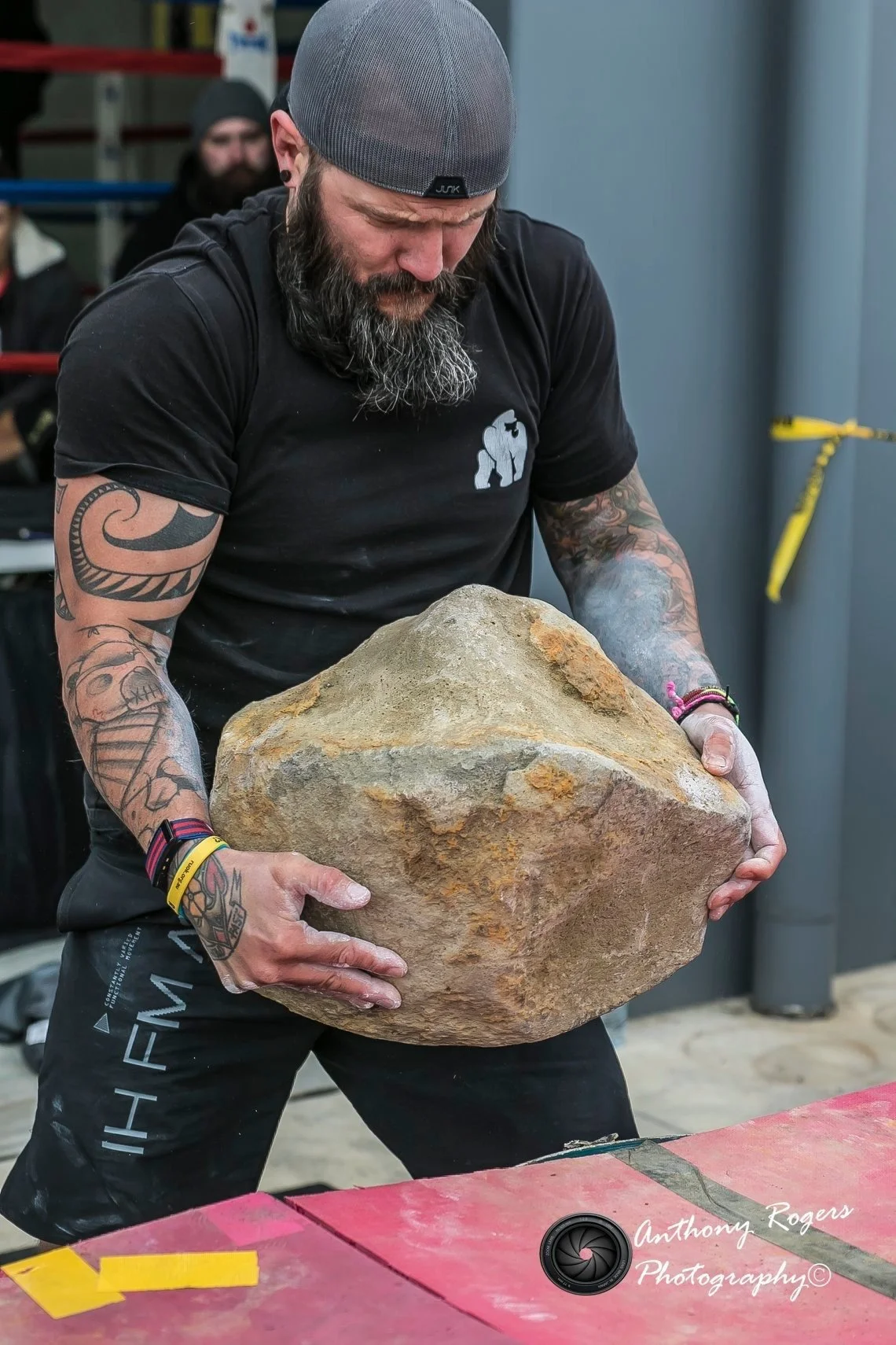 A man with tattoos wearing a gray cap and black shirt is lifting a large rock at a gym or fitness facility.