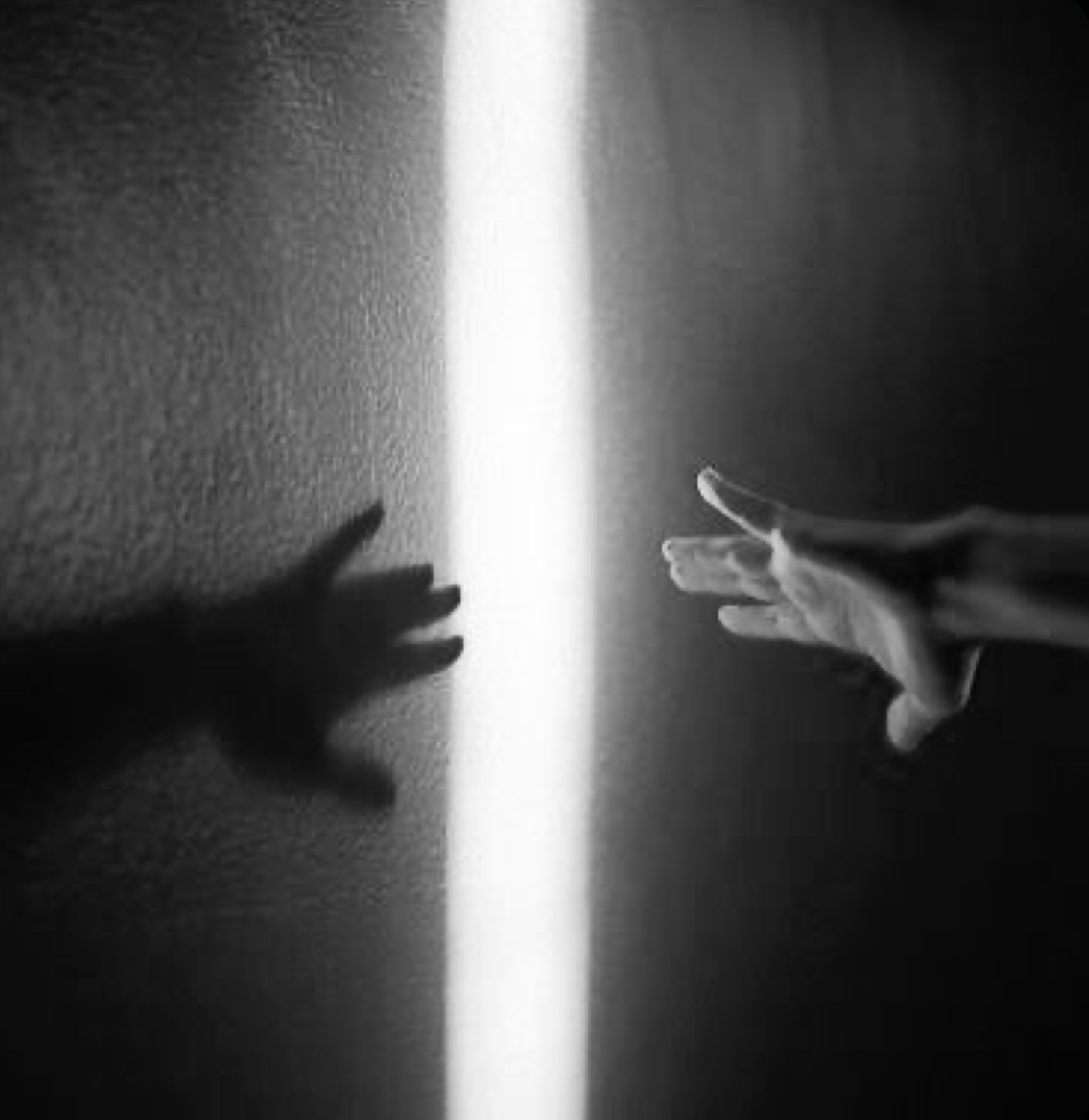 Two hands reaching towards each other, casting shadows on opposite walls, with a vertical beam of light separating them.
