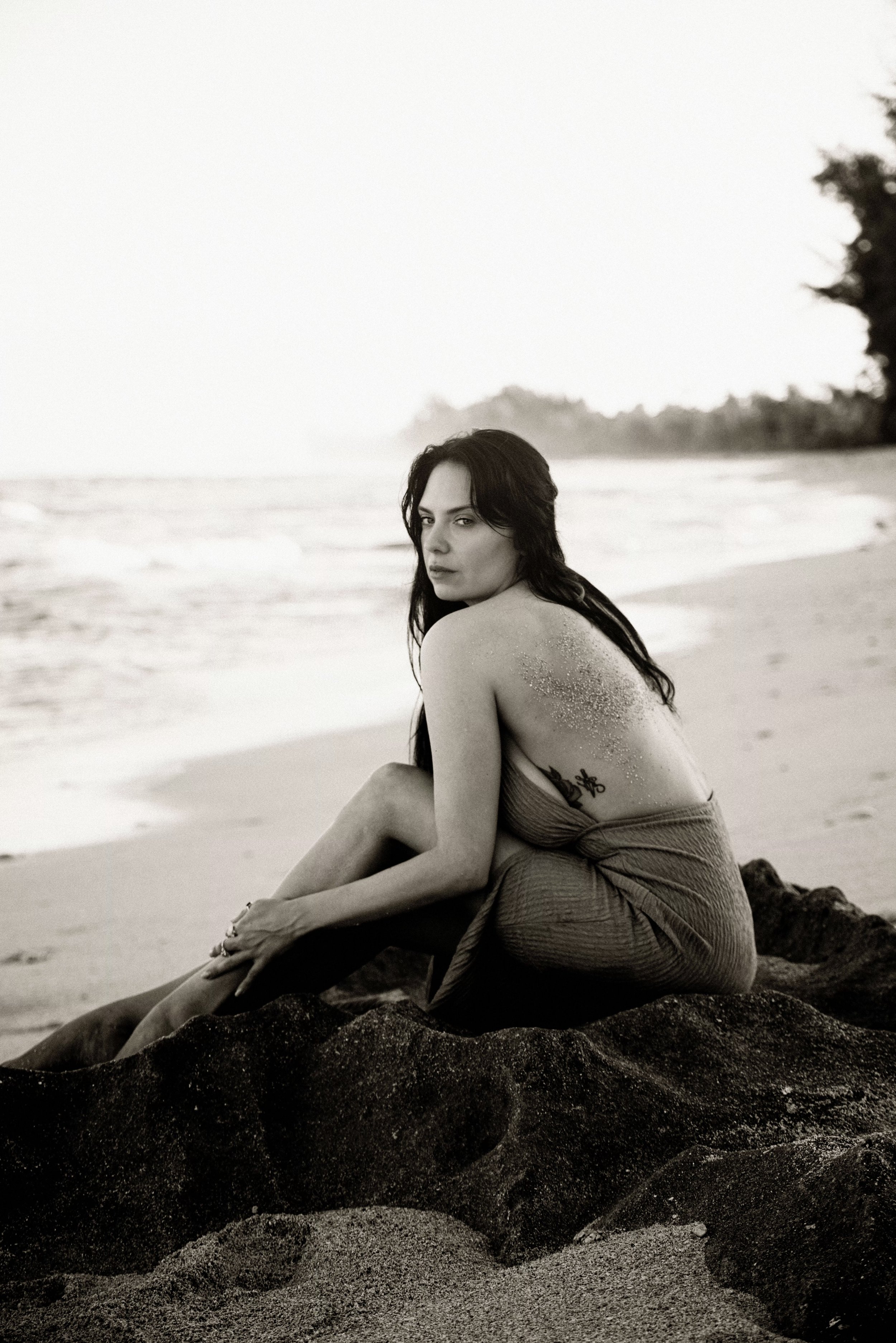 A woman with long dark hair sitting on a rock on the beach, looking at the camera, with sand on her shoulder, in a black and white photo.
