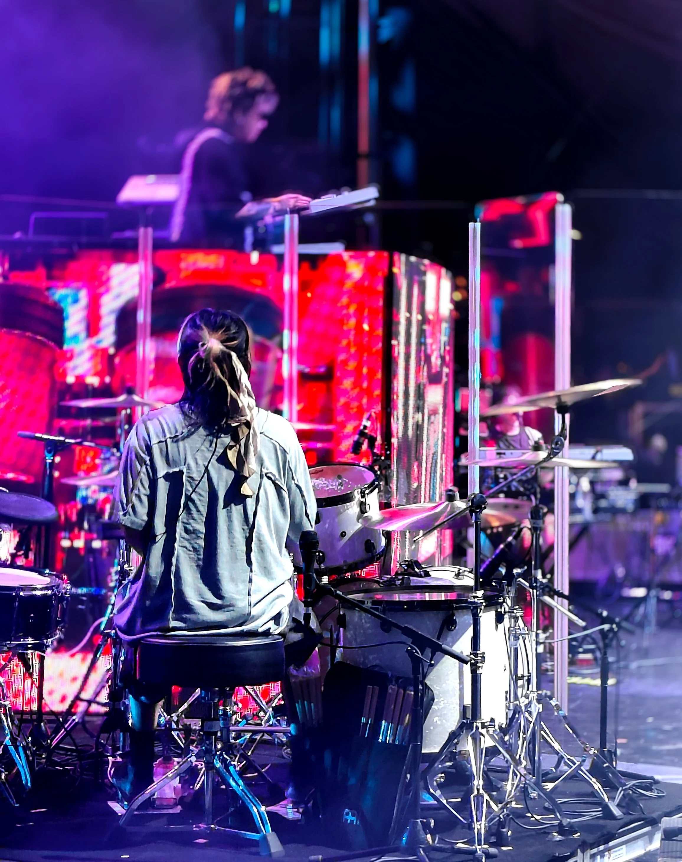 Musician playing drums on stage with a singer in the background, illuminated by colorful stage lights.