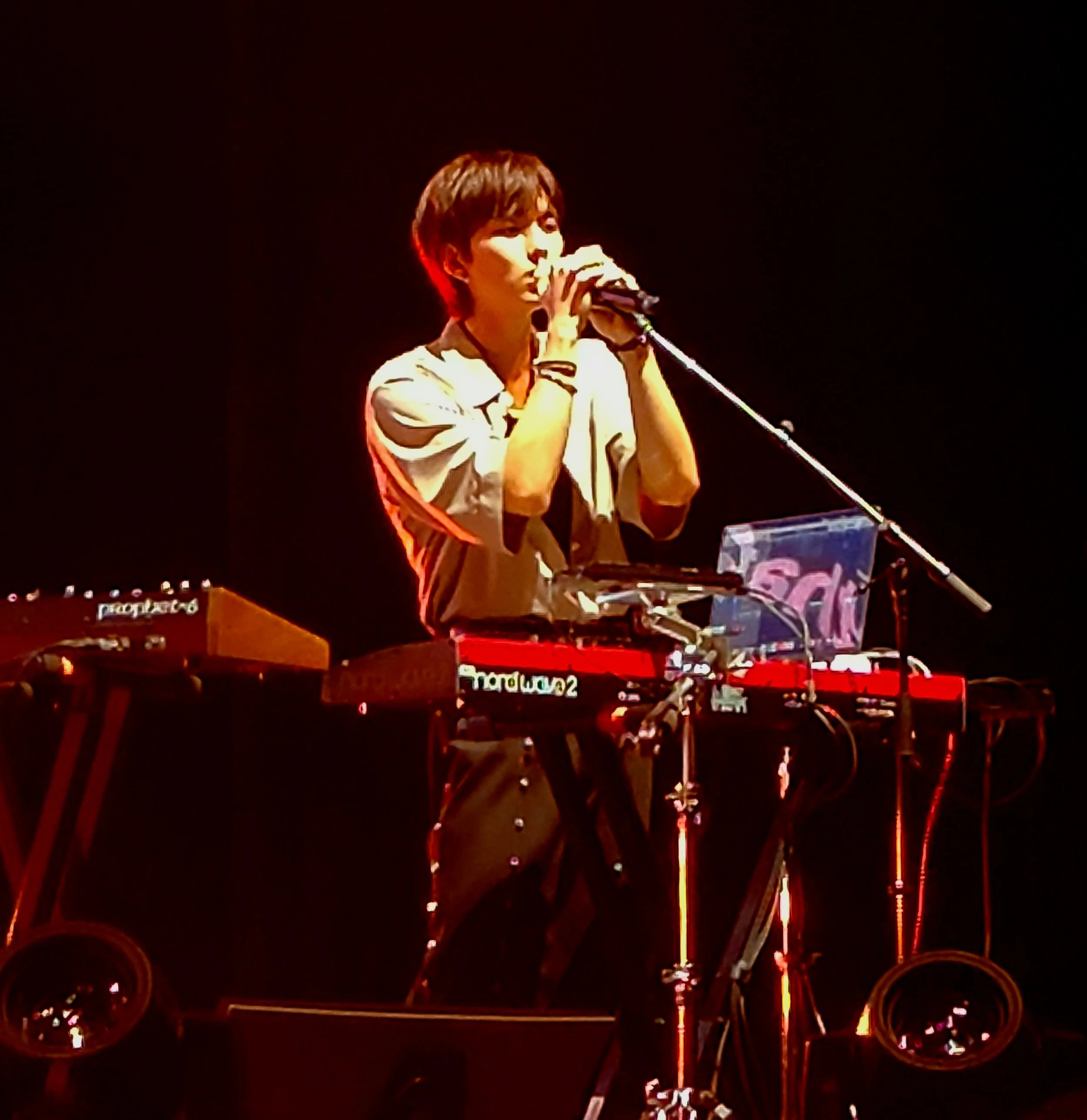 A musician standing behind multiple electronic music equipment on a stage, holding a microphone near their mouth, with a dark background and red stage lighting.