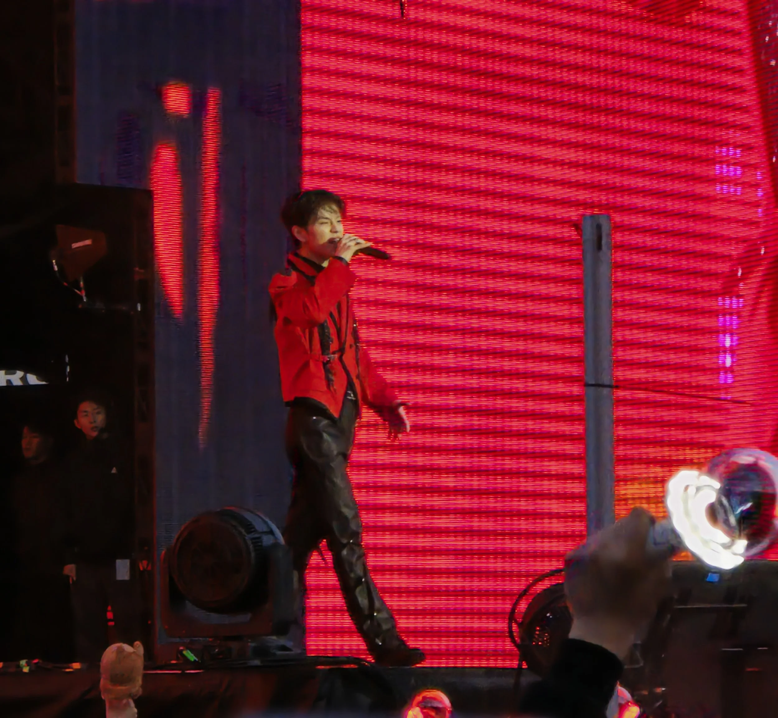 A performer singing into a microphone on stage with a red LED background, wearing a red jacket and black leather pants, during a concert.