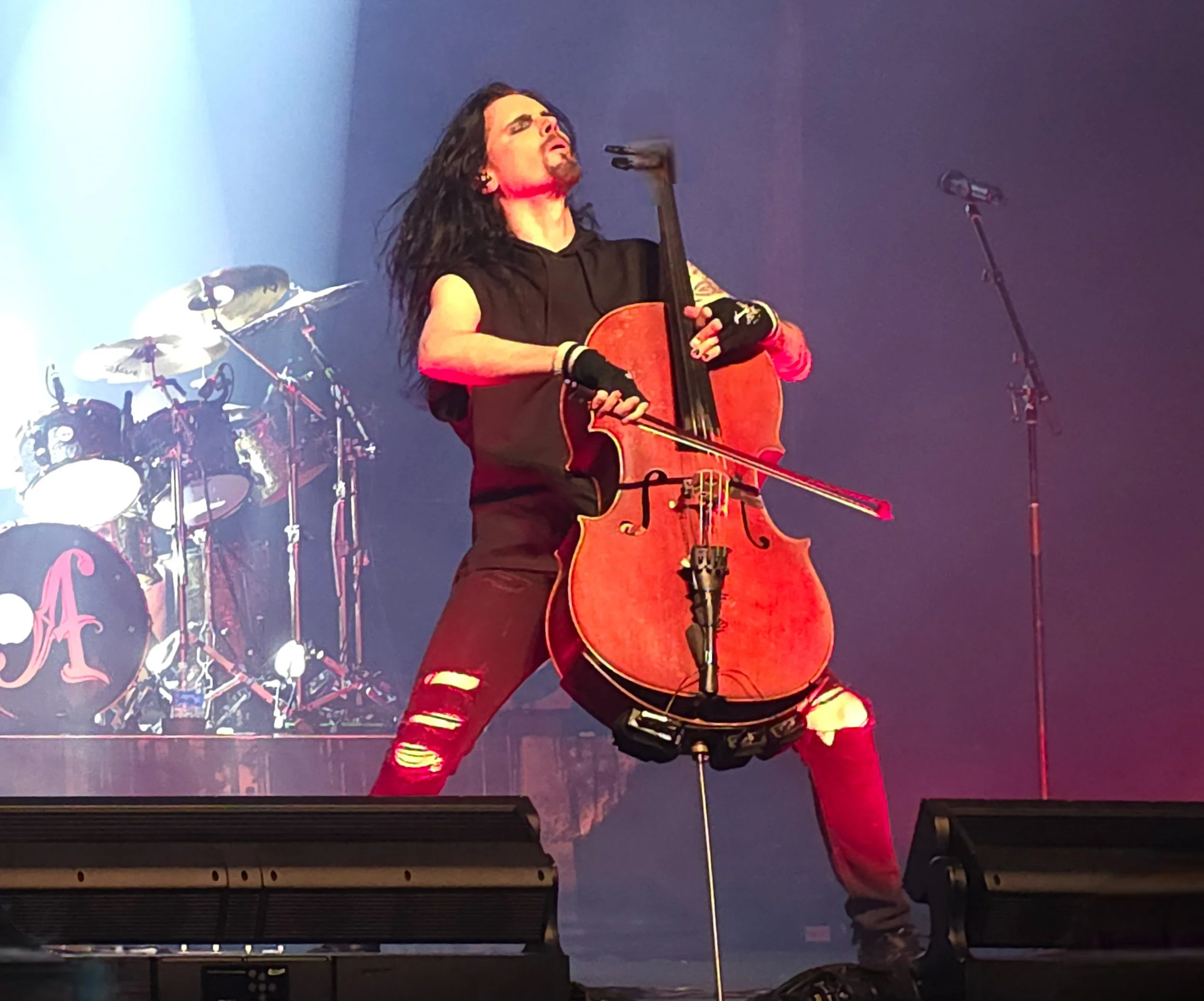 A musician with long black hair and tattoos playing an acoustic cello on stage, wearing black sleeveless top, ripped red jeans, and fingerless gloves, with a drum set in the background.