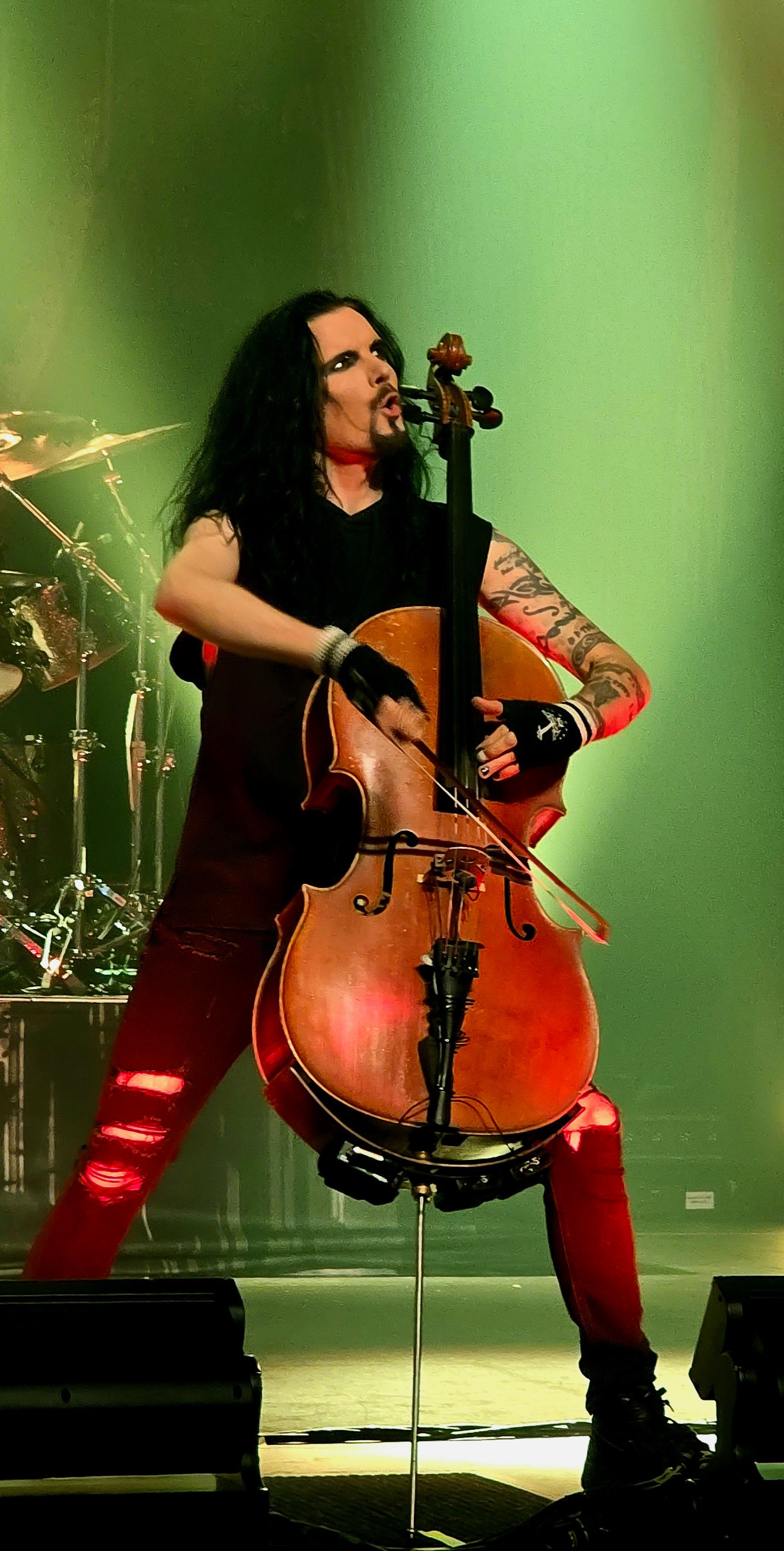 A male musician with long black hair, tattoos, and wearing red ripped jeans is playing a cello during a live performance with a green background.