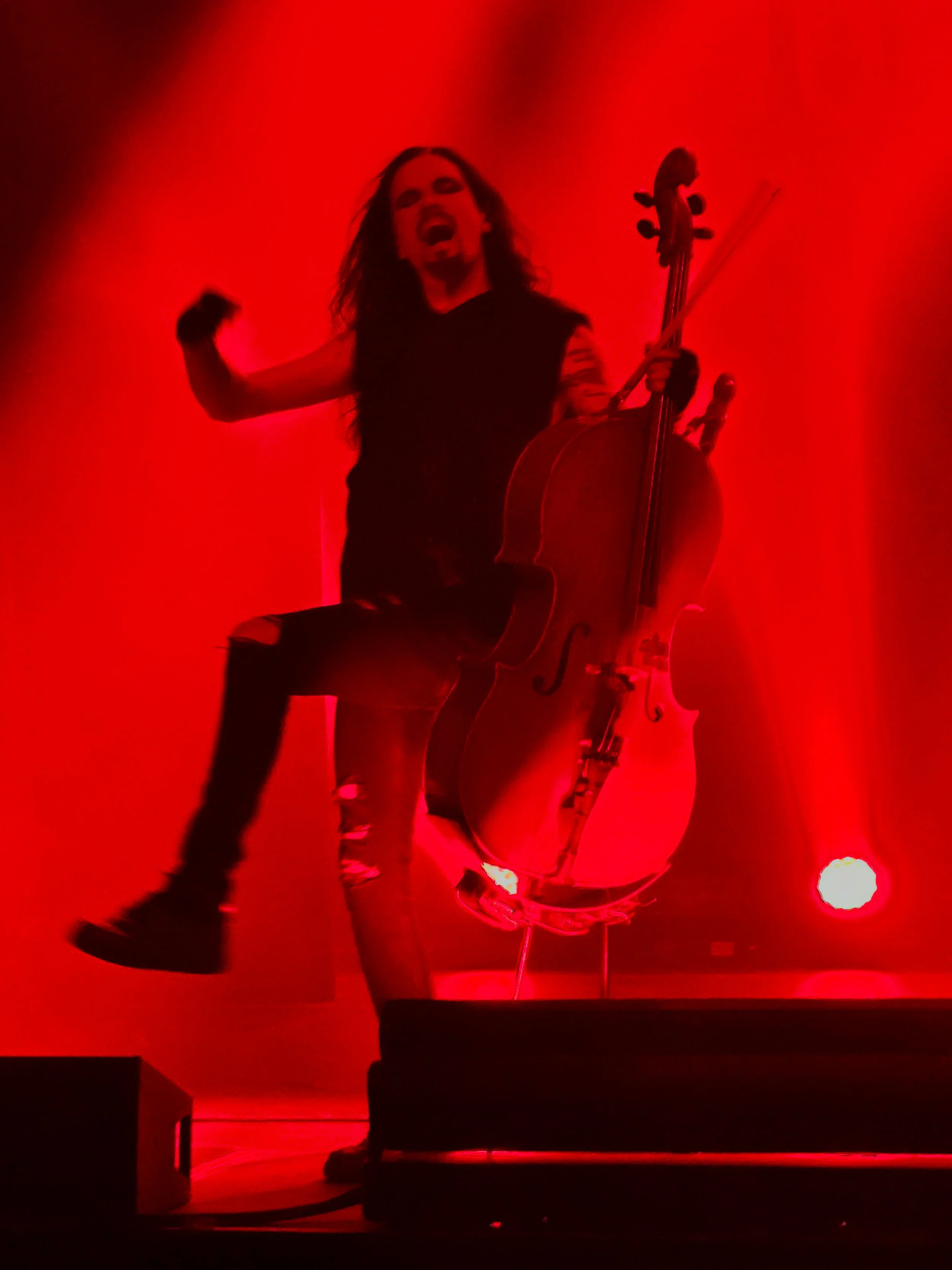 A musician with long hair and tattoos, playing a cello on stage illuminated by intense red lighting, with their mouth open in a powerful expression.