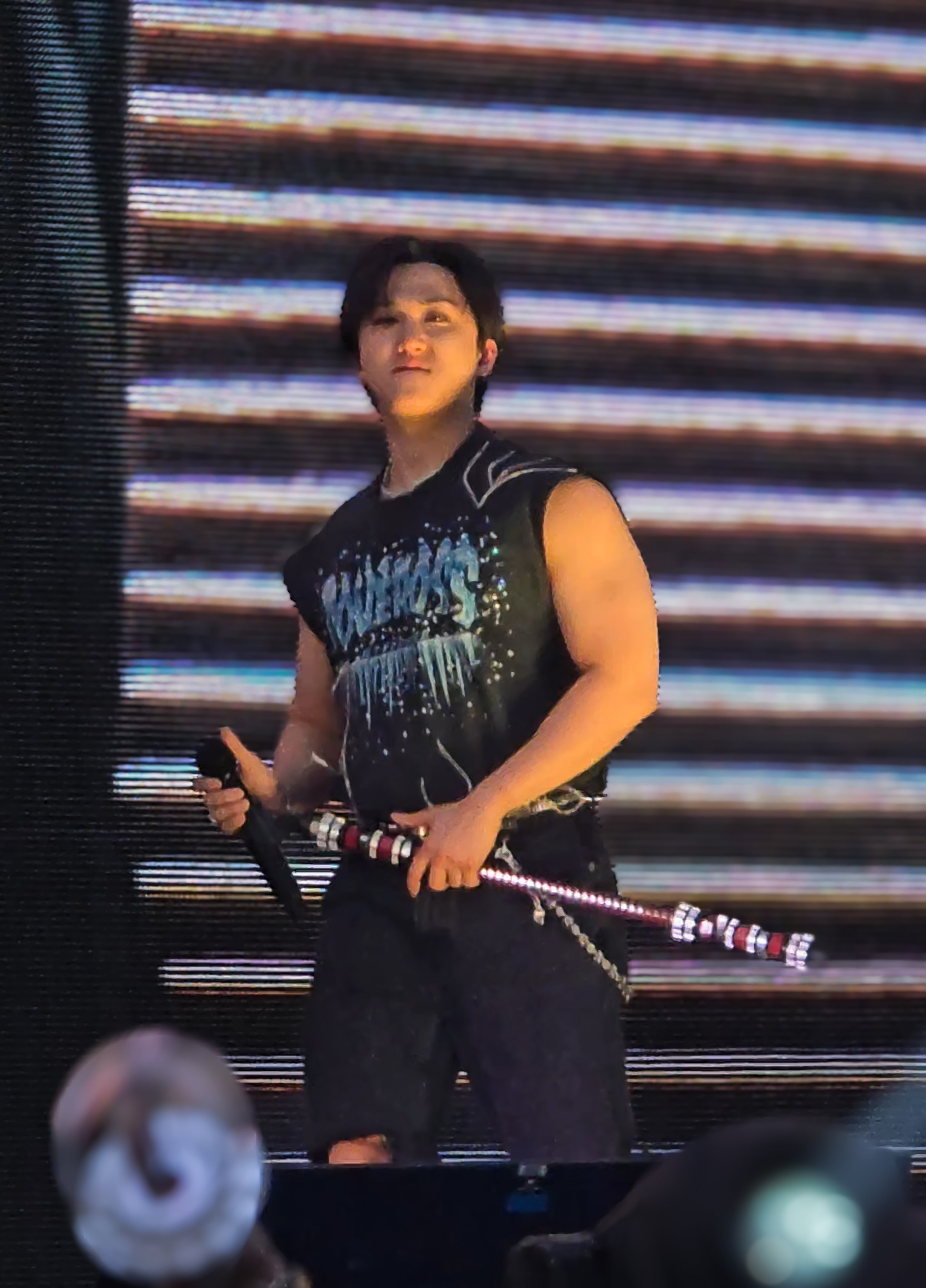 A young male performer on stage, holding a microphone and a staff with lights, wearing a sleeveless graphic T-shirt and dark pants, in front of a large LED screen with horizontal lines.