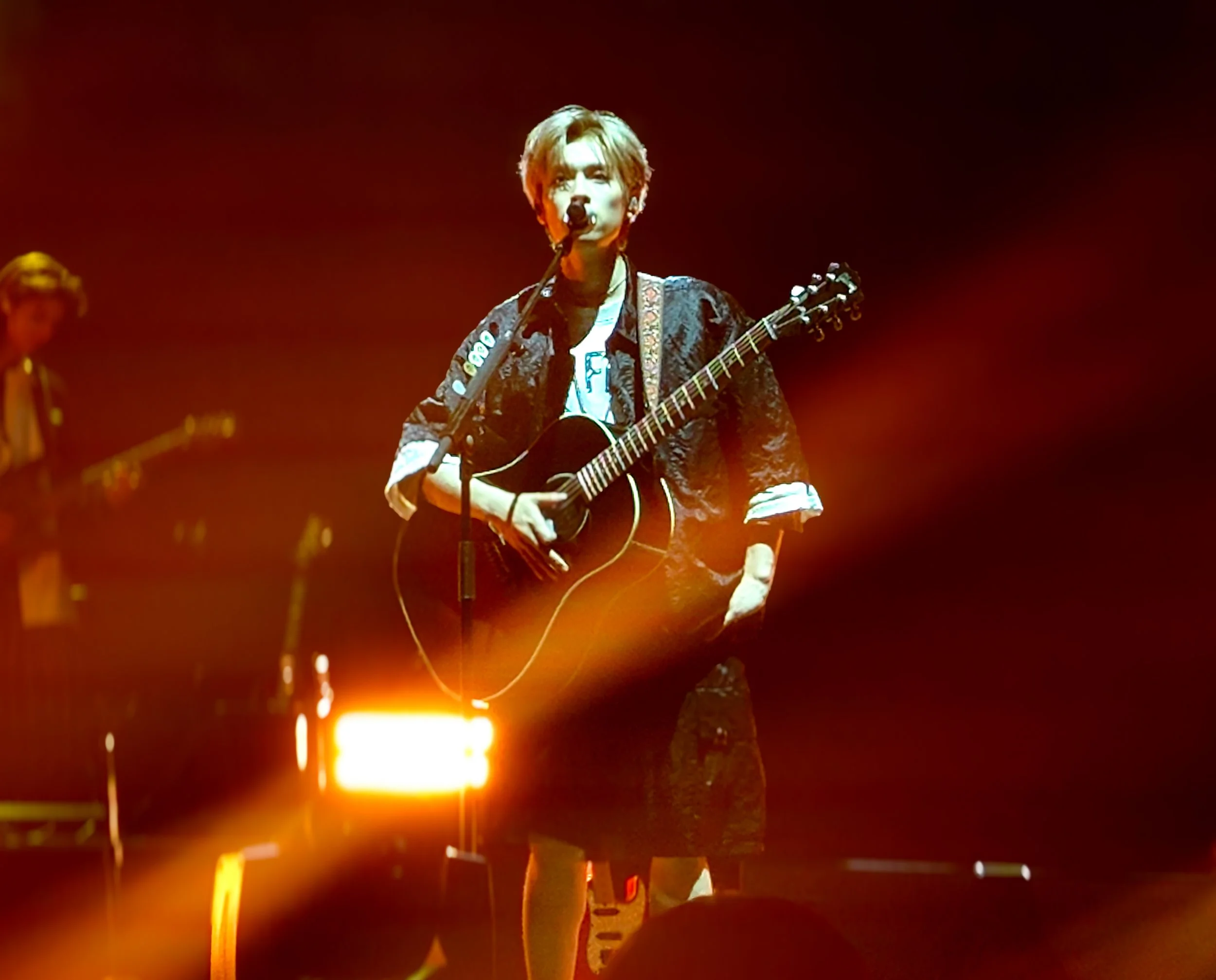 Musician with short hair playing an acoustic guitar and singing into a microphone on stage, lit with orange and yellow lights, with another musician in the background.