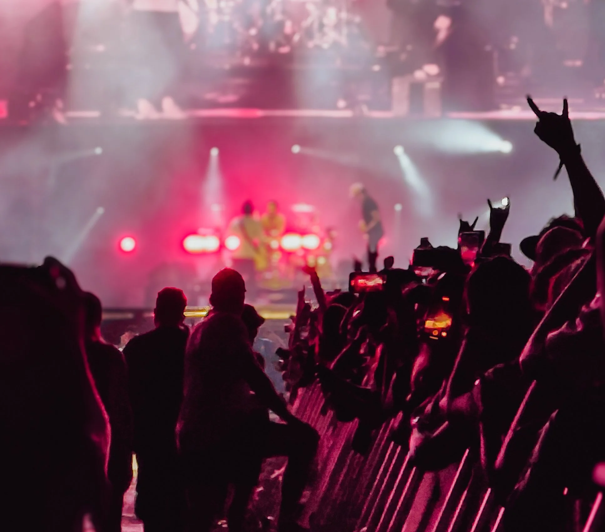 A concert scene with performers on stage illuminated by vibrant pink and red lights, and an audience in the foreground raising their hands and taking photos.