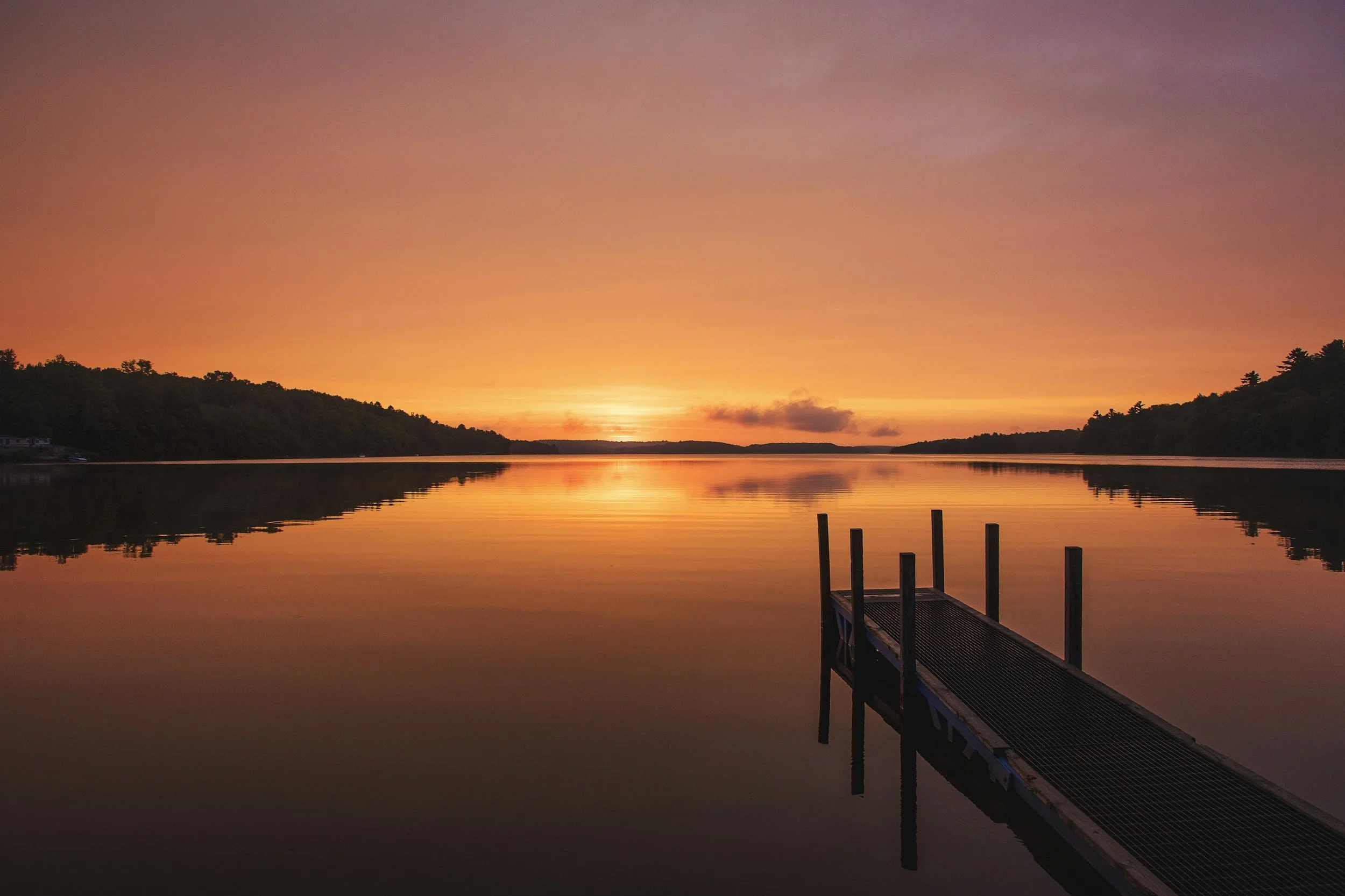 A calm lake during sunset or sunrise with a wooden dock extending into the water, and a colorful sky with hues of orange, purple, and pink reflected on the water.