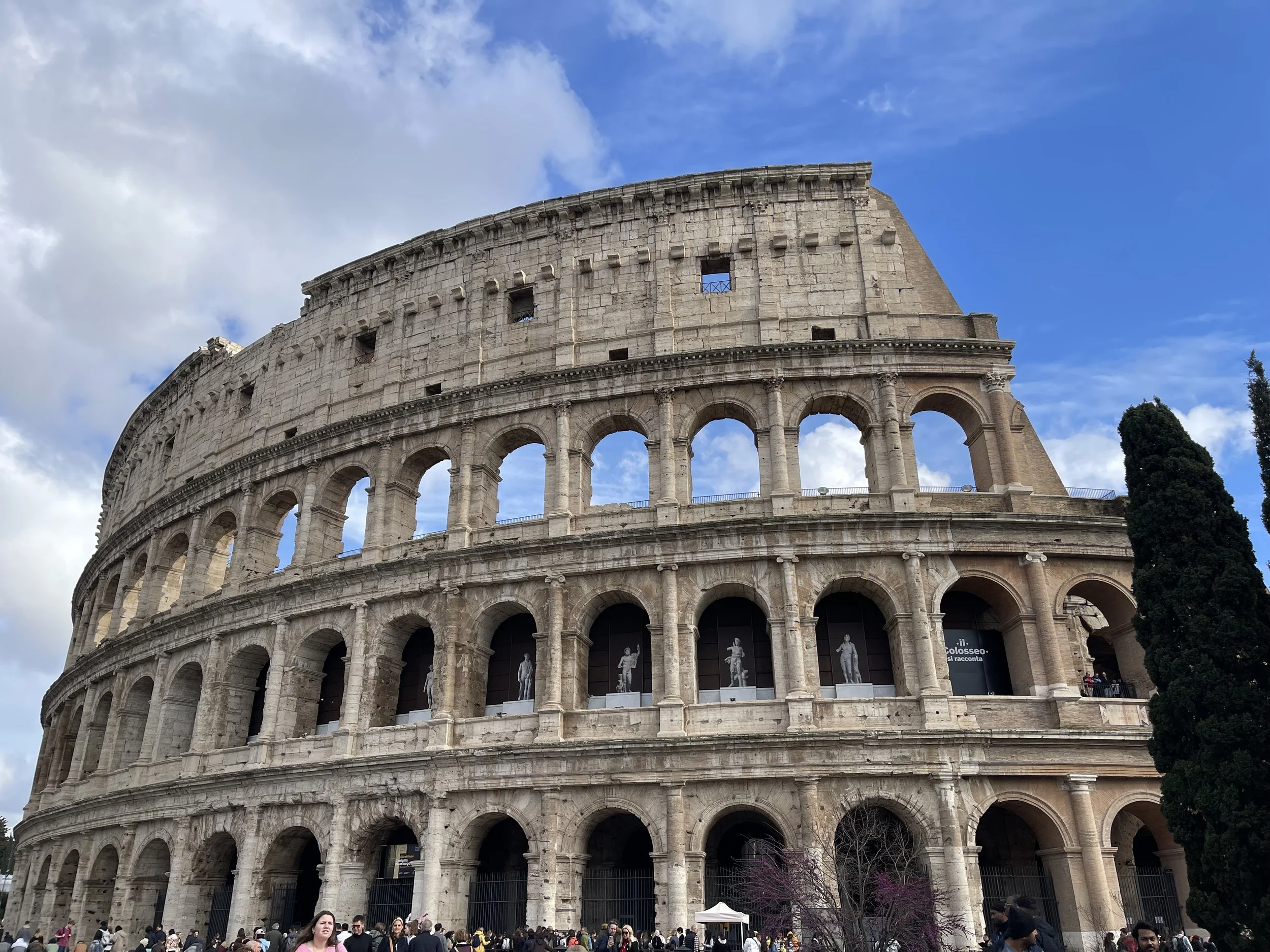 The Roman Colosseum, an ancient amphitheater with multiple arches, partly cloudy sky, and people gathered at its base.