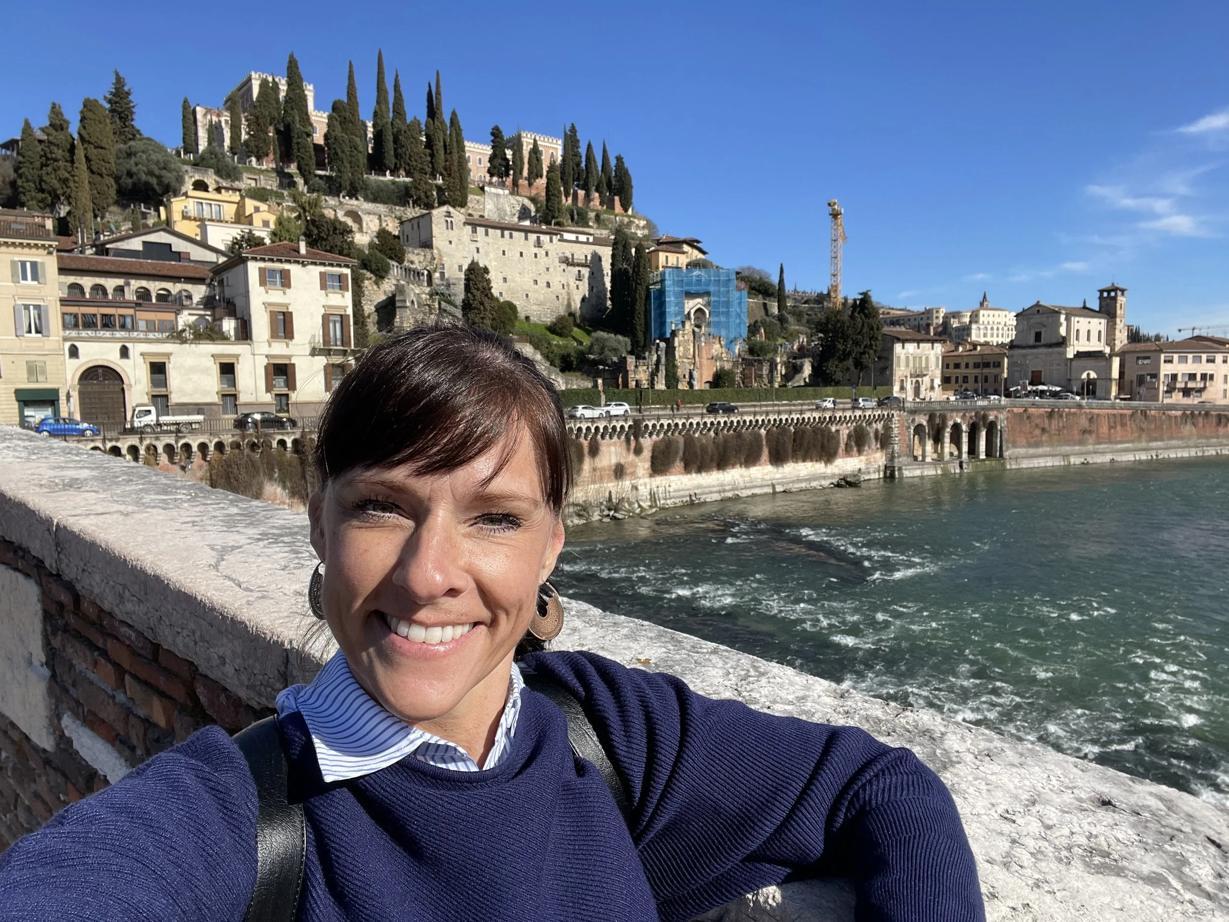 A woman taking a selfie by a canal, with historic buildings and a hillside with trees in the background on a sunny day.