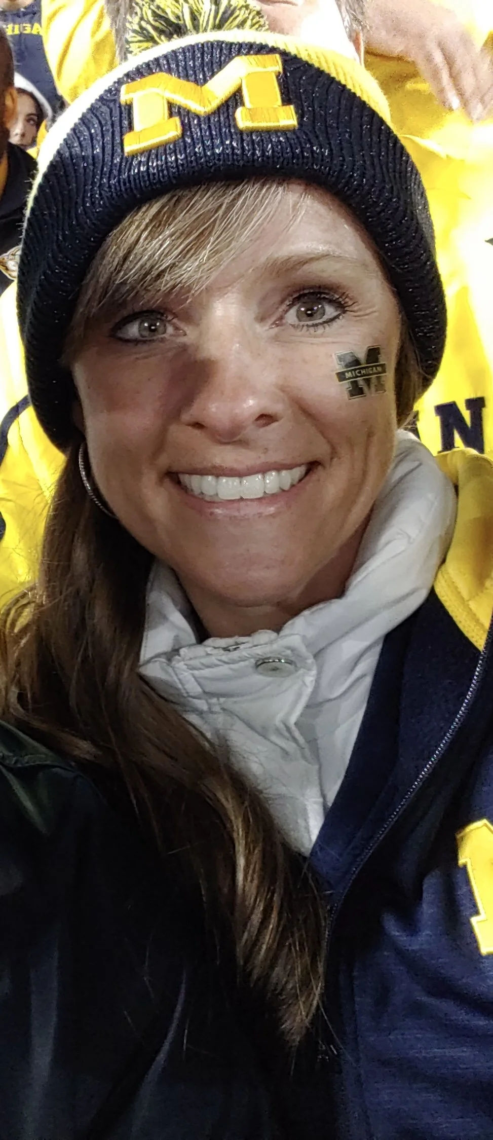 A woman wearing a black and yellow beanie with the letter 'M' on it, a white puffy jacket, and Michigan Wolverines stickers near her eye, smiling at a sports event in a crowd dressed in Michigan apparel.