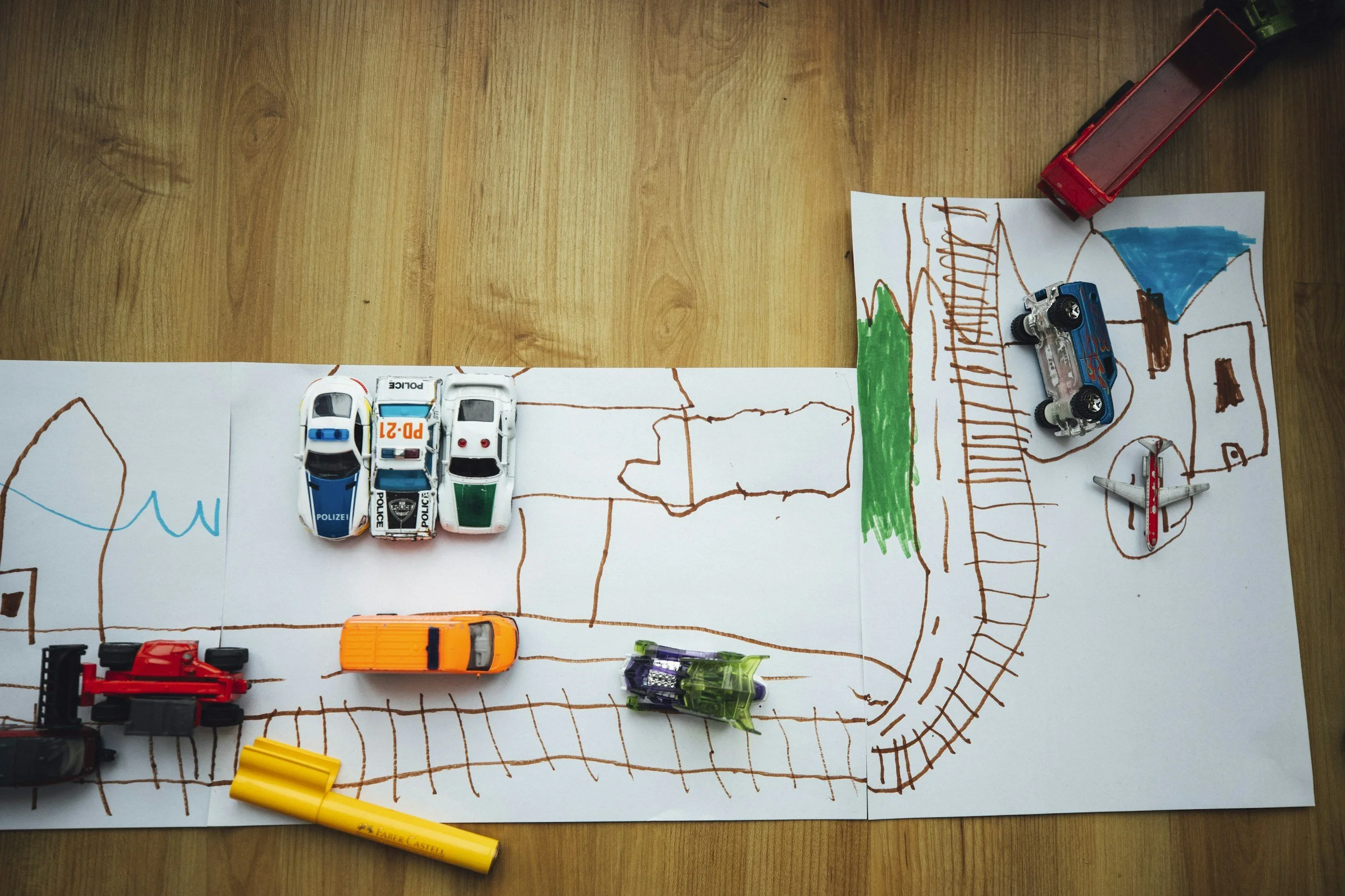 Children's drawing on large paper with toy cars, a truck, and a marker on a wooden surface. The drawing depicts a simple outdoor scene with roads, a house, trees, and a railroad, created with brown, green, blue, and red markers.