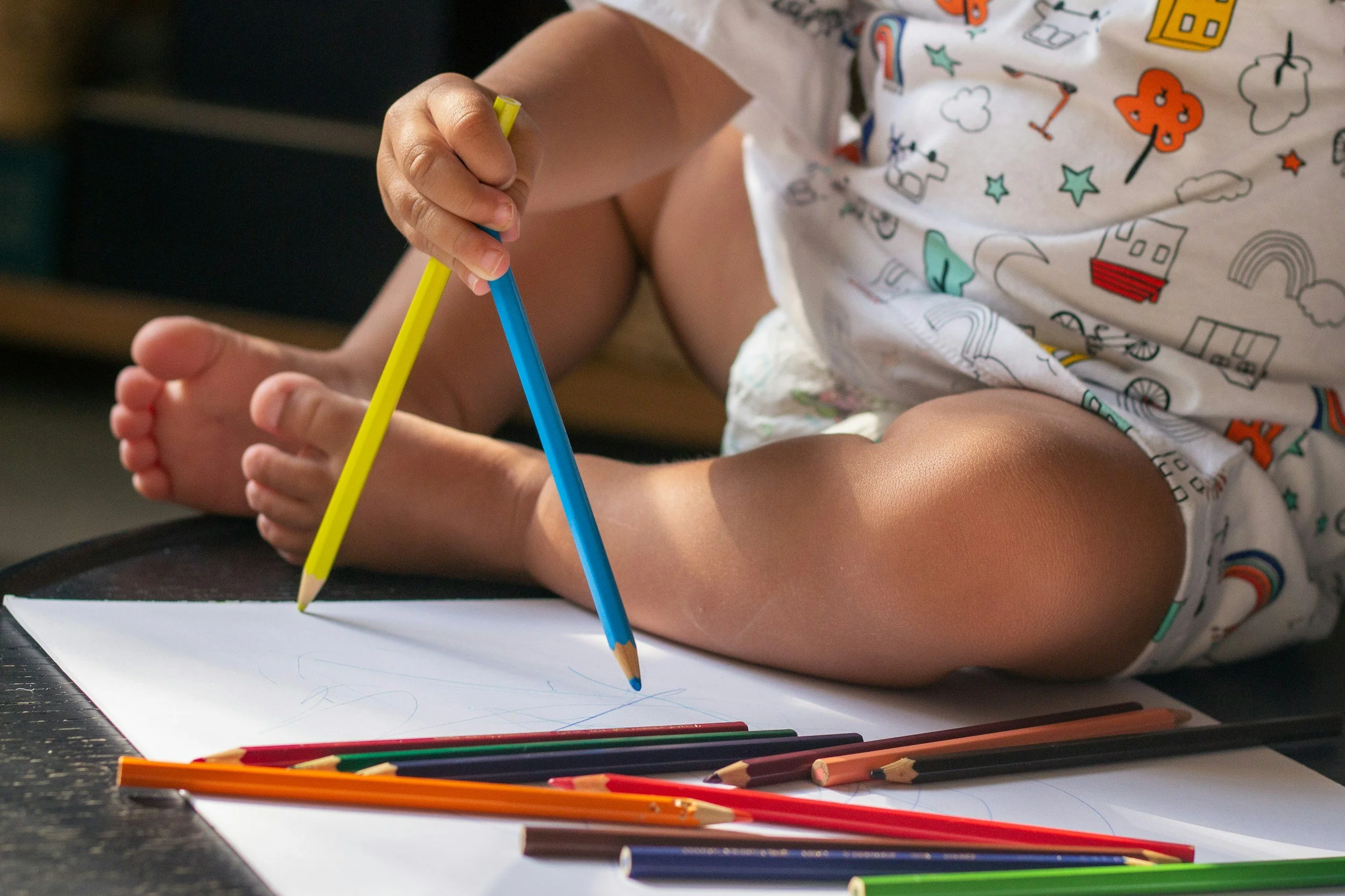 A young child sitting on a table, drawing on a large sheet of white paper with colored pencils, surrounded by more colored pencils, wearing a colorful, patterned shirt.