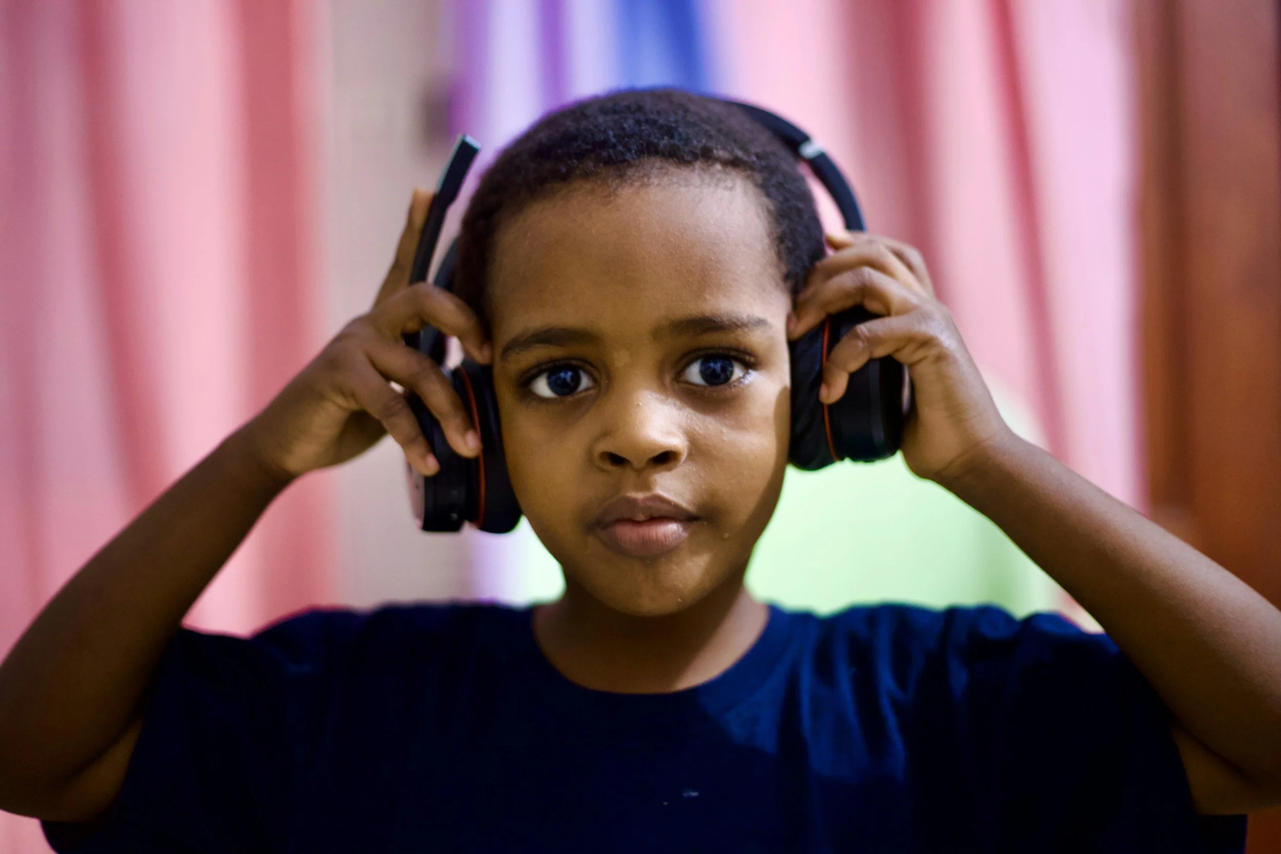 A young boy with dark hair and skin, wearing a black shirt, is holding headphones over his ears and looking directly at the camera. The background features pink and green curtains.