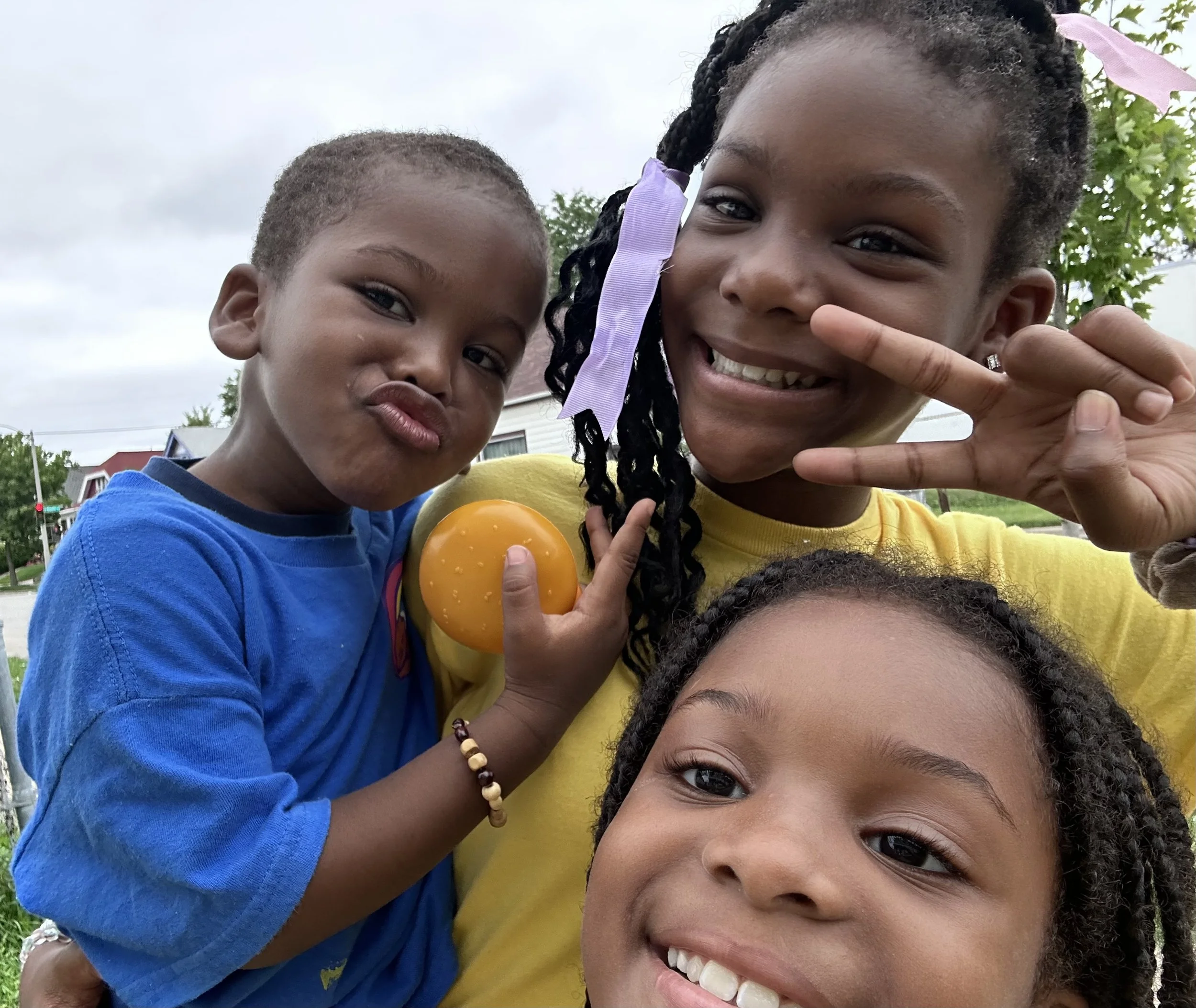 Three children smiling and posing outdoors on a cloudy day, one holding a yellow rubber ball, with a background of houses and trees.