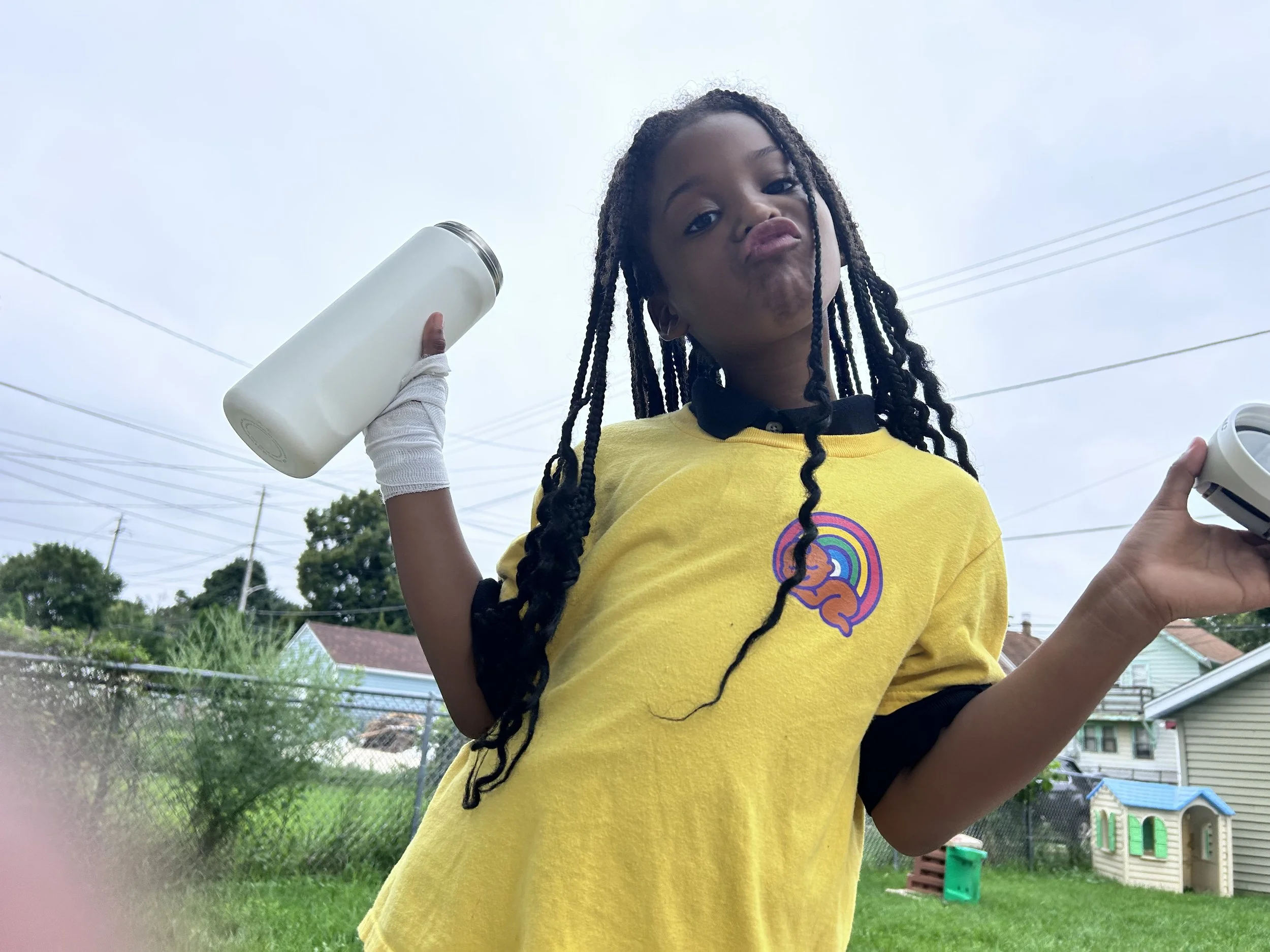Young girl with long braided hair wearing a yellow T-shirt making pouty face outdoors, holding a white water bottle in one hand and a small item in the other, with a suburban backyard background.