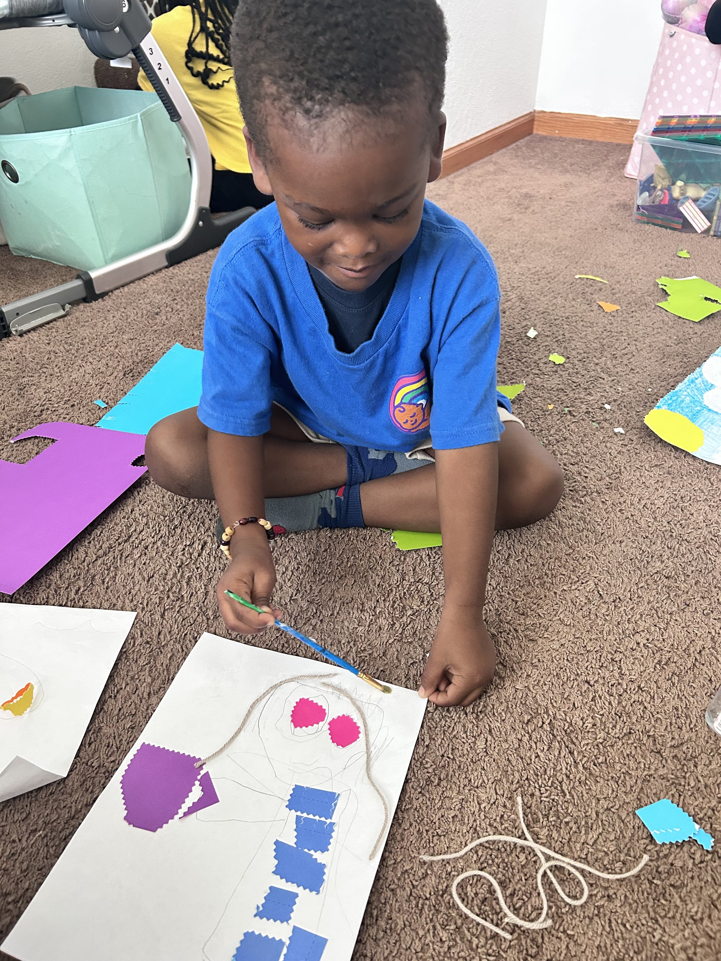 A young boy sitting cross-legged on carpeted floor, painting a craft picture with foam stickers, with colorful paper scraps and craft supplies around him.
