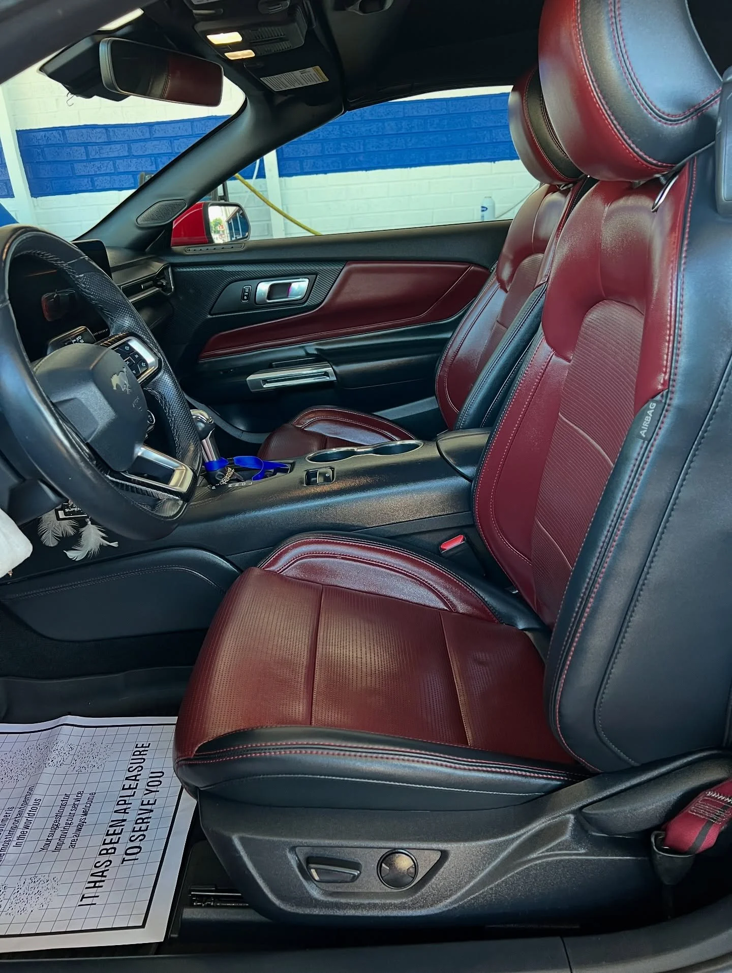 Interior of a vehicle featuring red and black leather seats, a steering wheel, dashboard, and side door panel. A paper floor mat is visible on the driver's side.