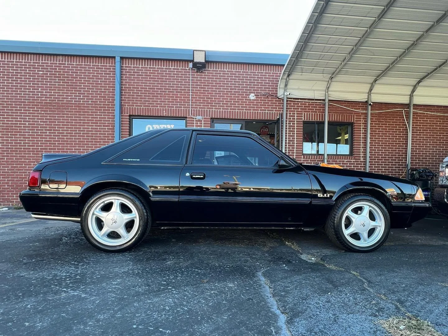 Black vintage Ford Mustang parked in front of a brick building with a blue roof and a covered awning.
