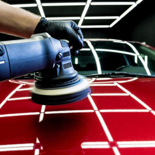 A person wearing a black glove polishes a red car with a handheld electric buffer in a garage with bright overhead lighting.
