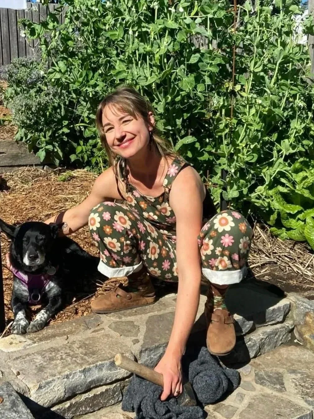 A woman in floral clothing crouching next to a black dog on a stone path in a garden with lush green plants.