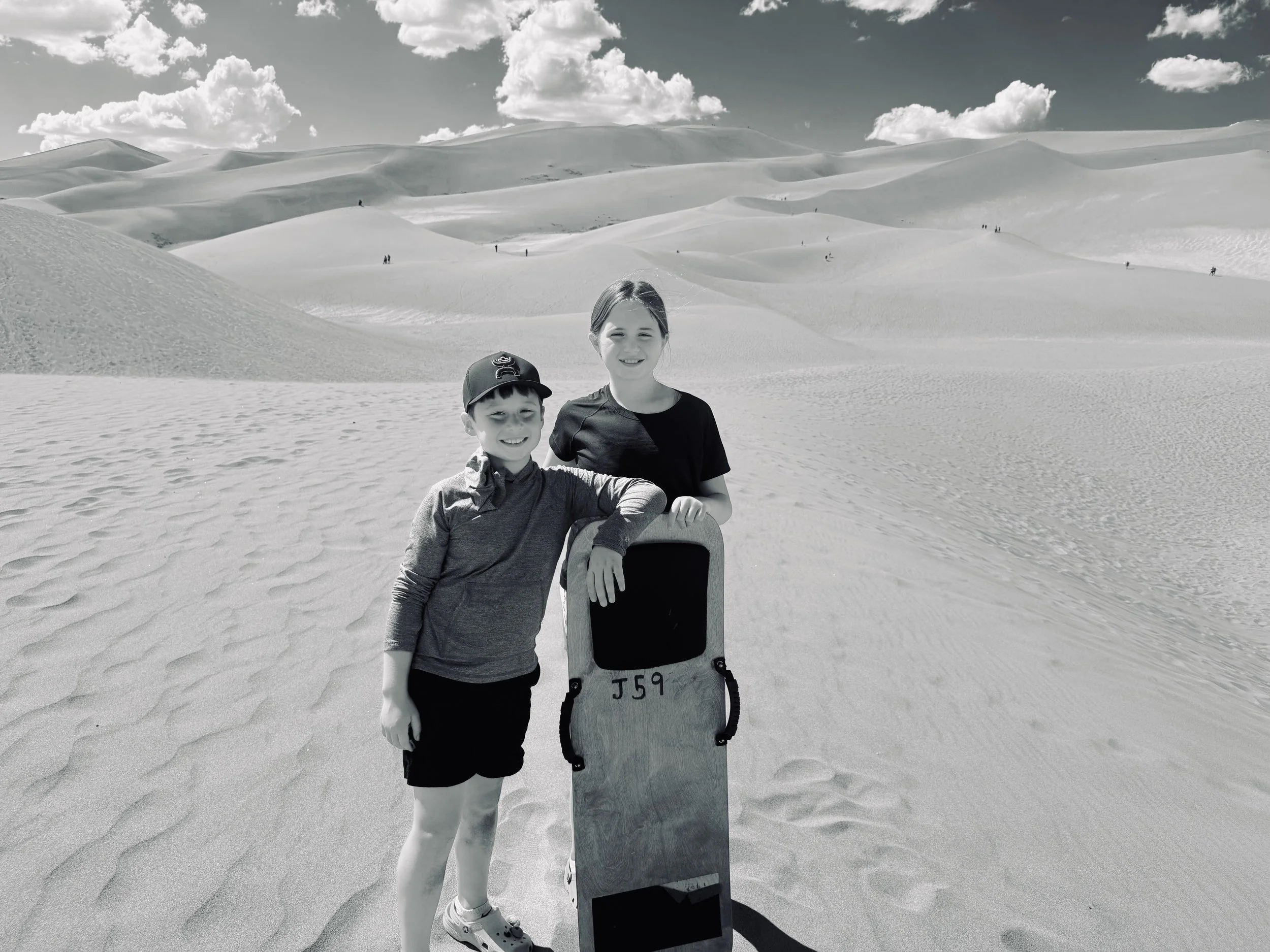 Two children, a boy and a girl, standing in a desert landscape with sand dunes under a partly cloudy sky, holding a skateboard.