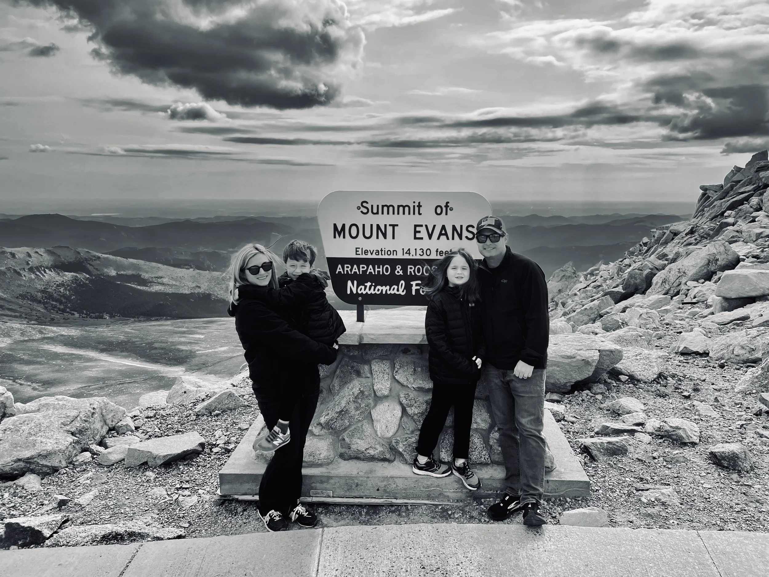 A family of four standing and smiling at the summit of Mount Evans, Colorado, with a large sign that reads ‘Summit of Mount Evans, Elevation 14,130 feet, Arapaho & Rockies National Forest’. The landscape features mountains and valleys in the background, with a cloudy sky overhead.