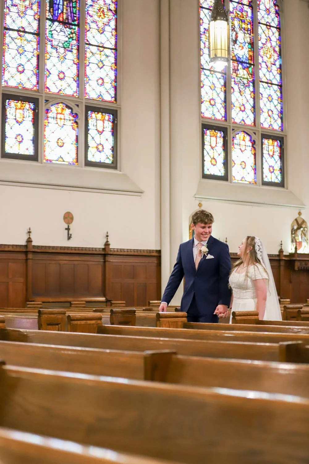 A newlywed couple walking hand in hand inside a church with stained glass windows in the background.