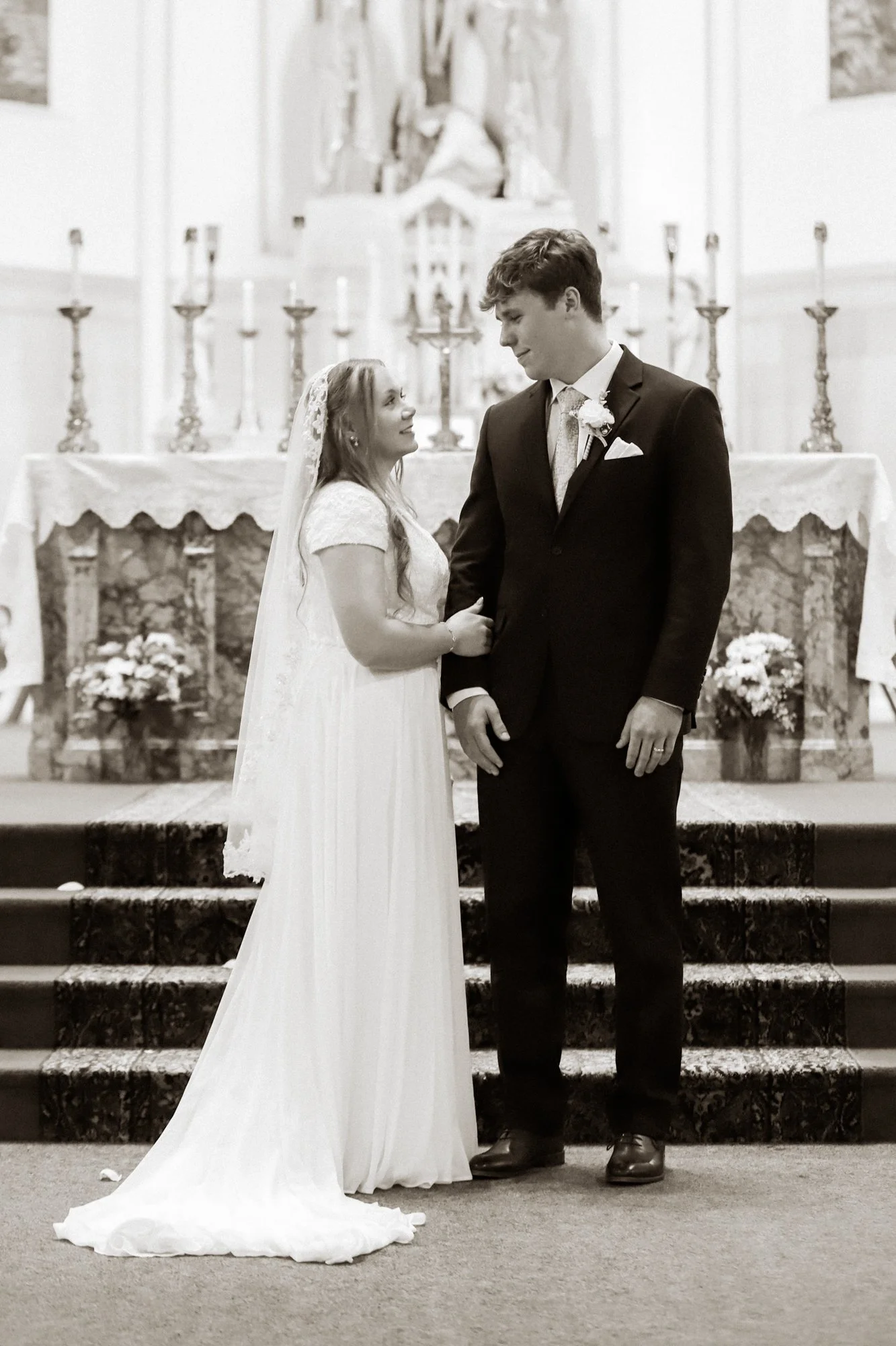 A bride and groom standing inside a church, facing each other and holding hands. The bride wears a white dress and veil, and the groom is in a dark tuxedo with a boutonniere. Behind them is an altar with candles and a crucifix.
