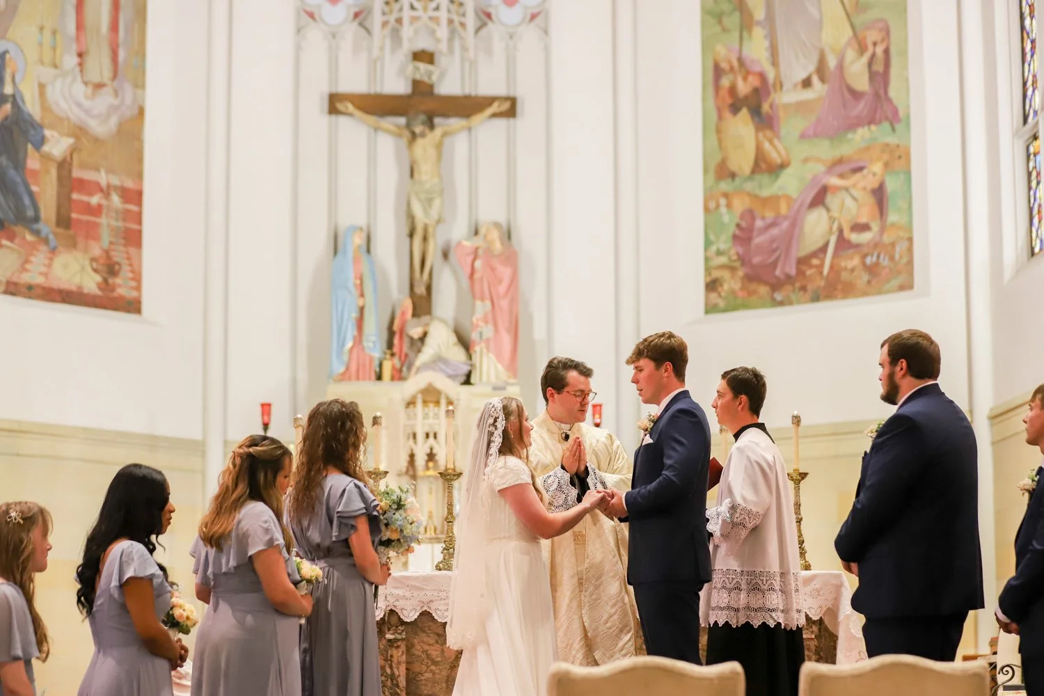 A wedding ceremony inside a church with a couple holding hands and praying, surrounded by bridesmaids and groomsmen, with religious artwork and a crucifix in the background.