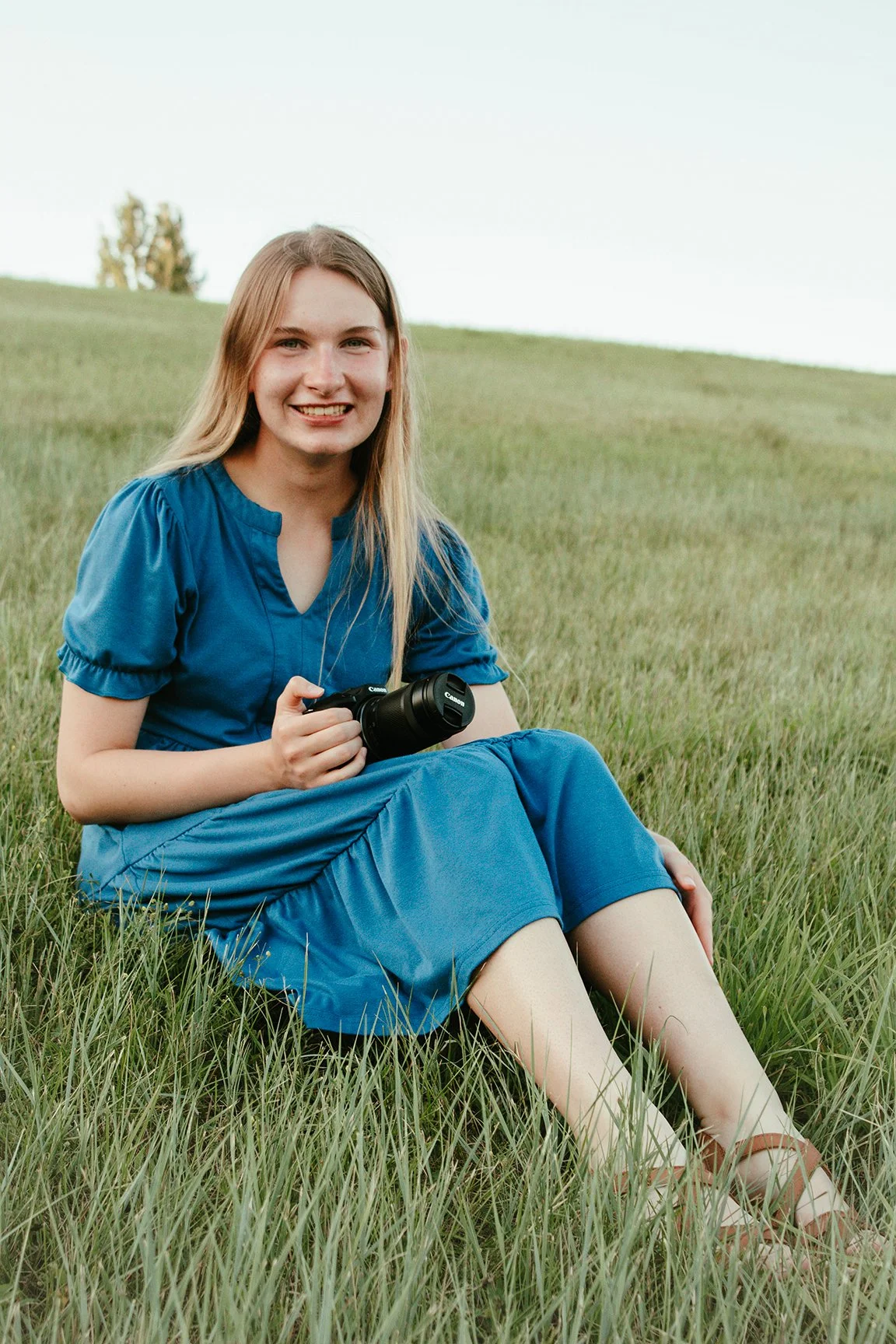 A young woman sitting on green grass in a field, holding a camera, wearing a blue dress, smiling.