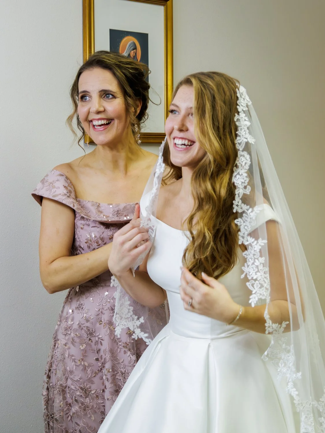 A joyful bride with a lace veil getting ready for her wedding, standing with a woman in a mauve floral dress who is helping her with her veil, behind them a framed religious artwork on the wall.