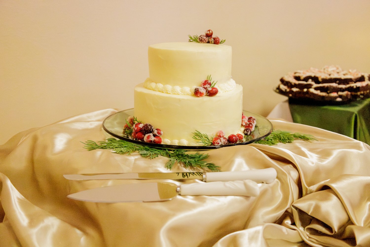 Two-tier white cake decorated with red berries and green herb-like accents, placed on a glass cake stand, with a gold satin tablecloth underneath.
