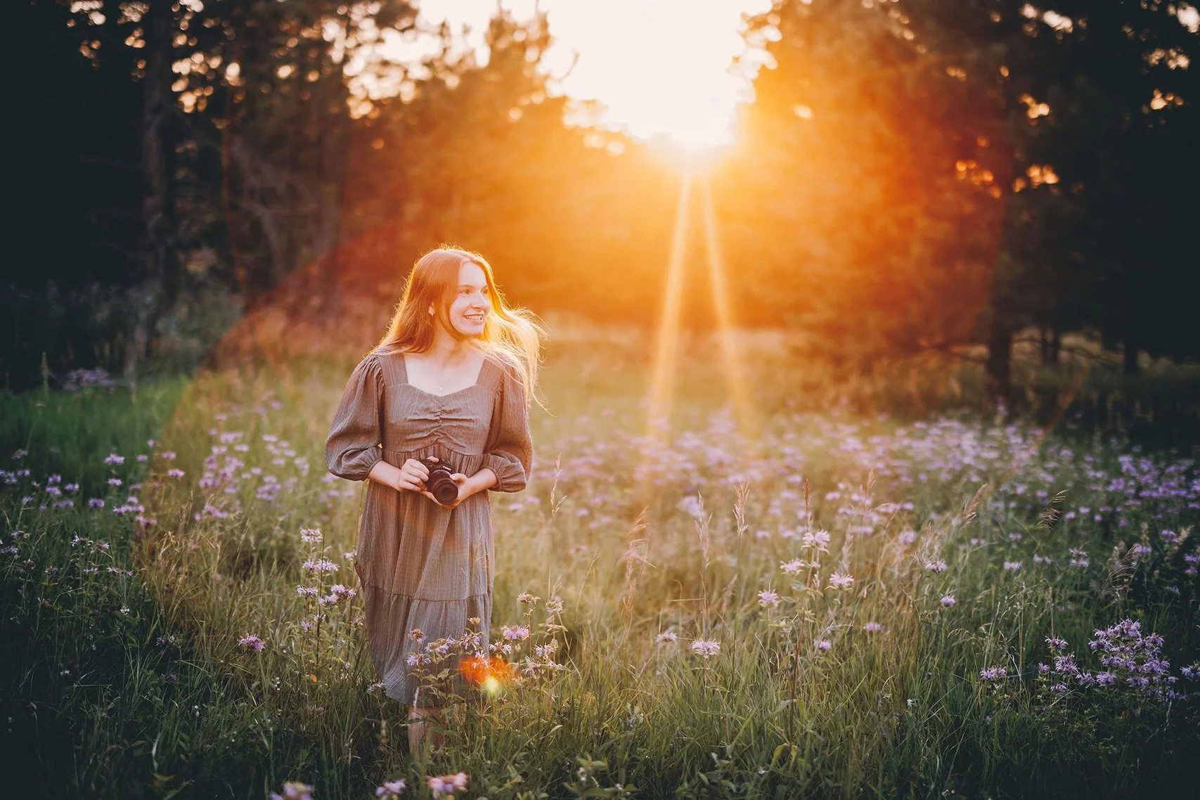 A young woman with long hair, smiling, holding a camera, standing in a field of purple flowers during sunset with trees in the background.