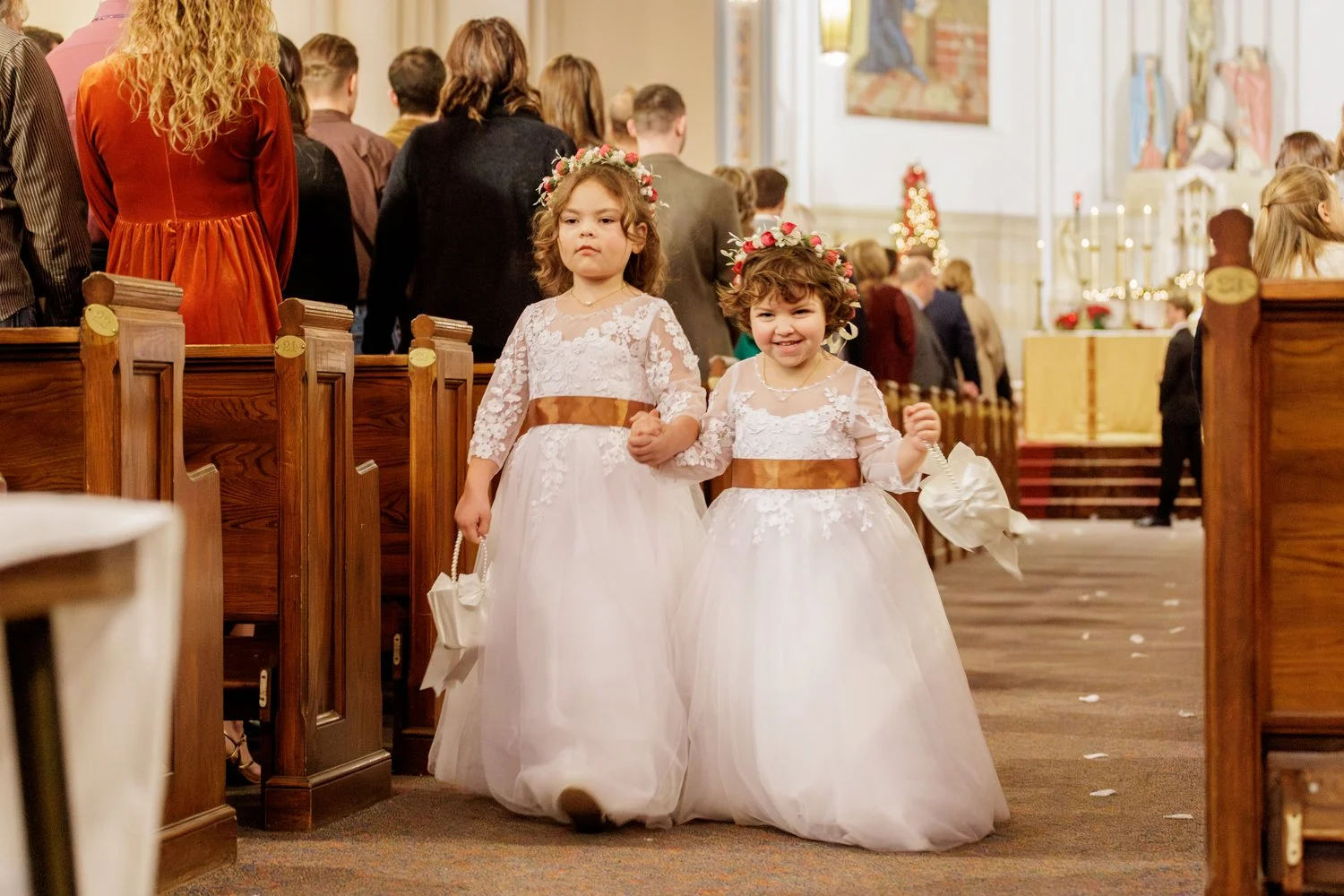 Two young girls dressed in white lace dresses with floral crowns walk hand-in-hand inside a church during a wedding ceremony while throwing flower petals on the ground.