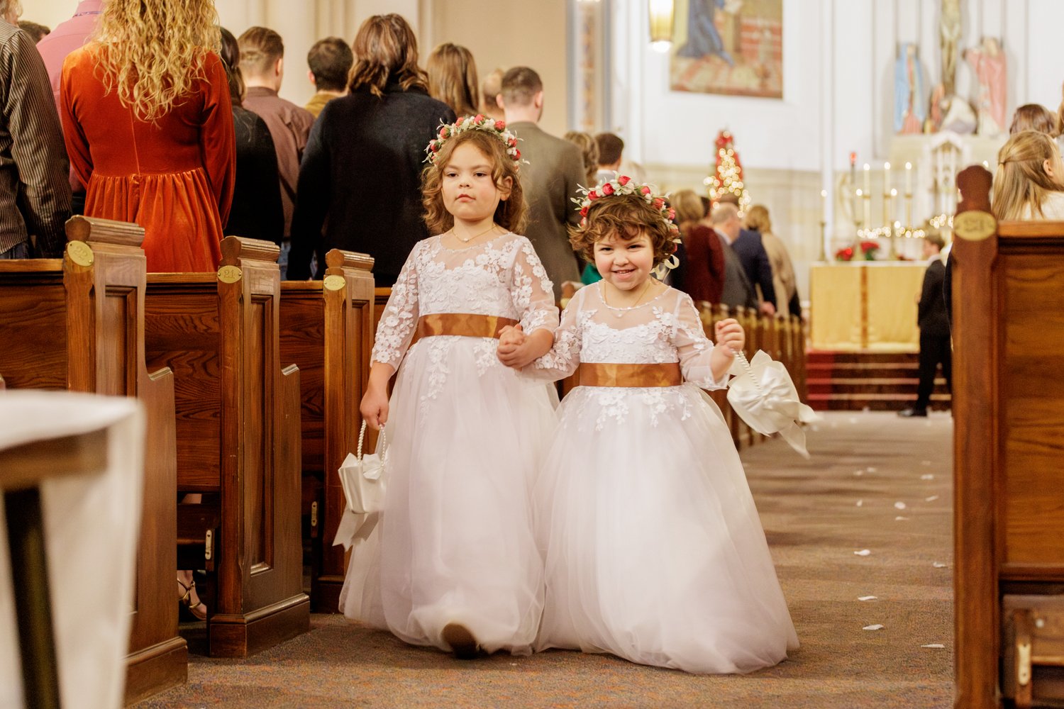 Two young girls in white lace dresses with brown sashes, floral headbands, and holding small white baskets, walking down the aisle of a church during a wedding ceremony and throwing flower petals on the ground.