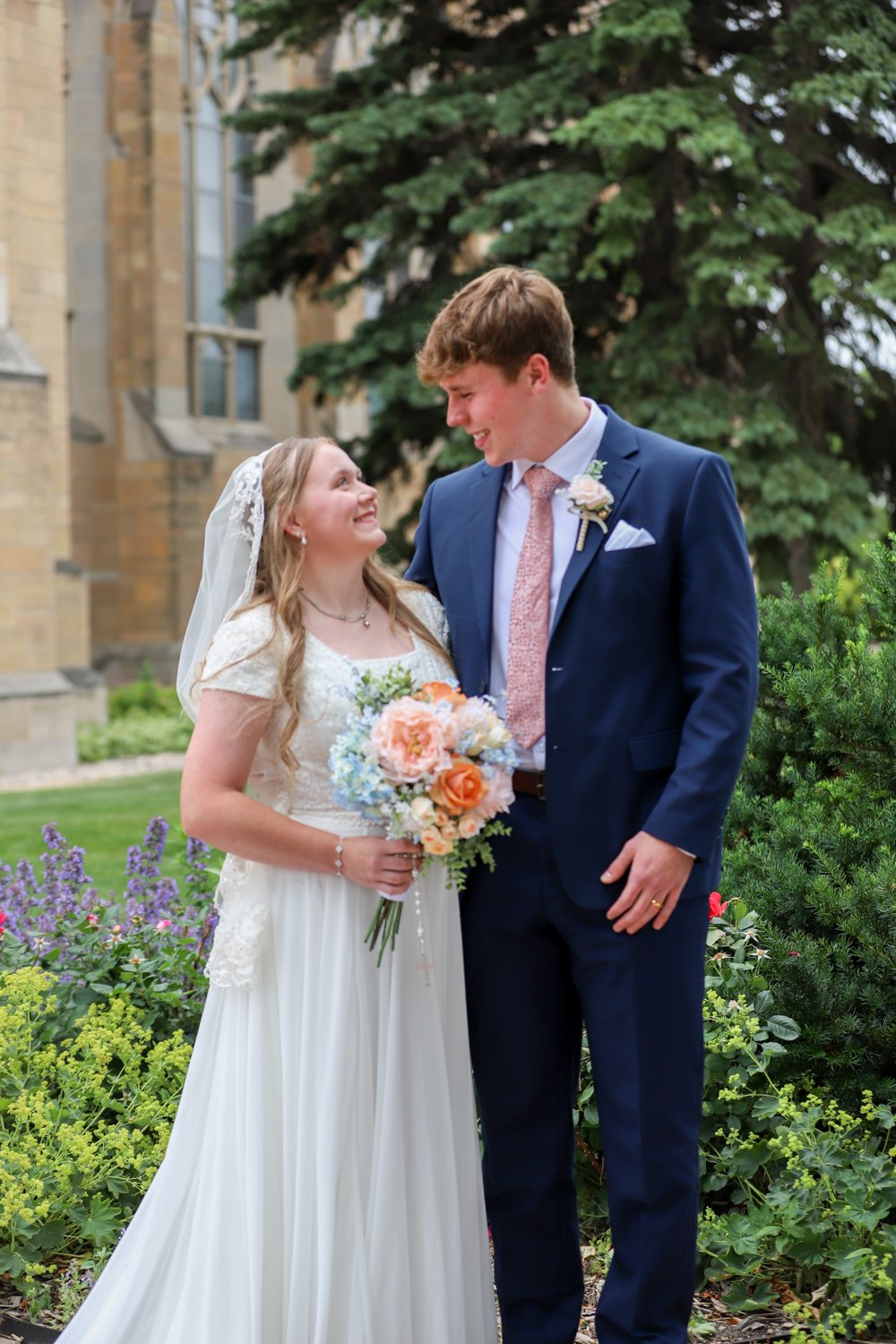 A bride and groom looking at each other and smiling outdoors, surrounded by greenery and flowers, with a church in the background.
