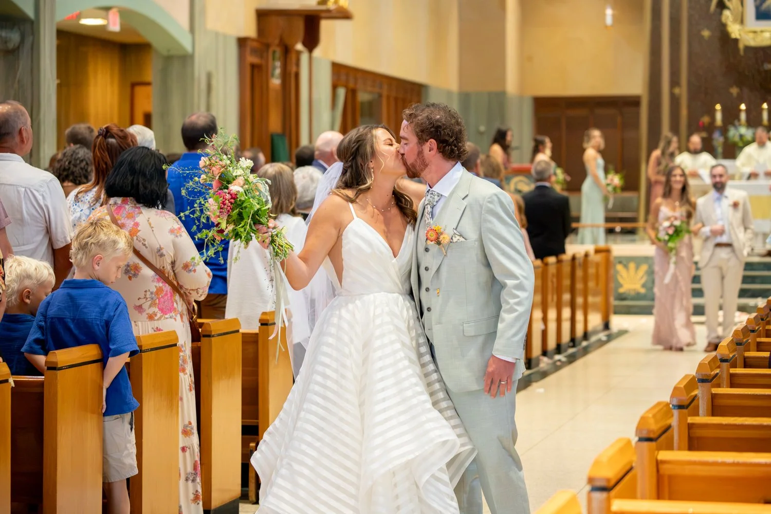 A bride and groom kiss at their wedding ceremony inside a Cathedral. The bride holds a bouquet of flowers, and guests stand in pews watching.