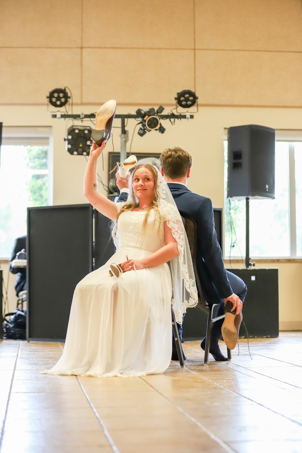 A bride and groom participating in a shoe game at their wedding reception, sitting back to back, each holding one of the other's shoes.