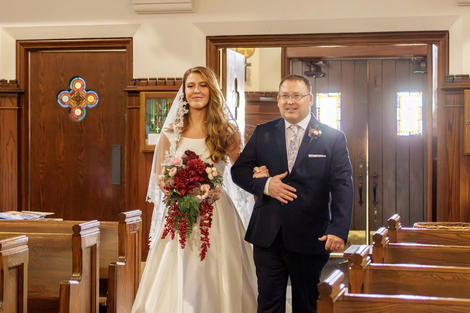 A bride in a white wedding dress holding a bouquet, walking down the aisle with her father inside a church with wood-paneled walls and stained glass windows.
